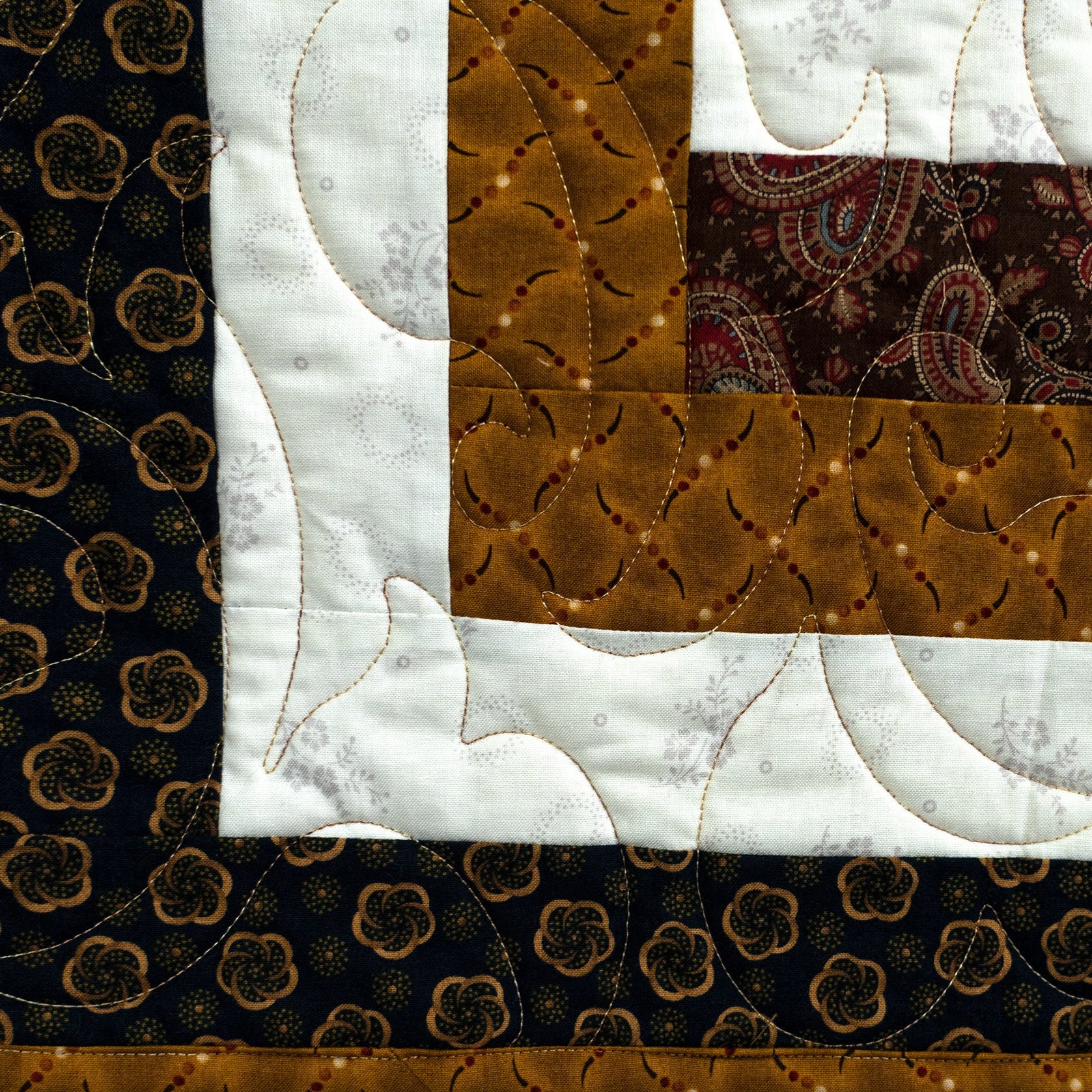 A close-up of a quilted fabric design with a geometric patchwork layout. The pattern features a dark brown and black floral border, a central white quilted section with wavy lines, a brown patterned section, and a dark paisley block.