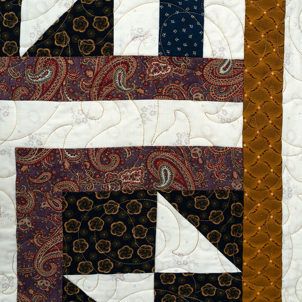 A close-up of a quilted textile with geometric and paisley patterned fabric blocks, including white curved quilting, dark navy floral sections, rust brown patterned areas, and a vertical rust-brown stripe.