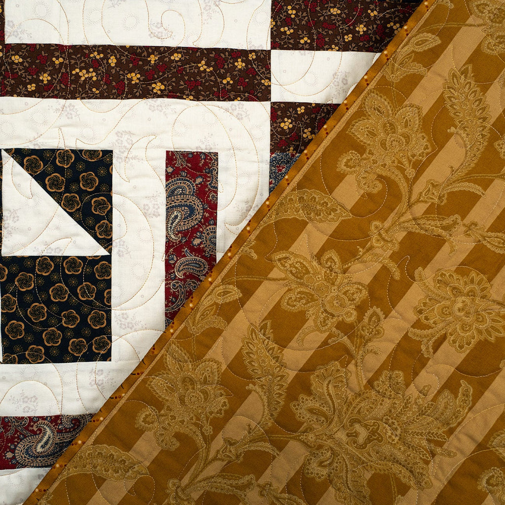 A close-up of a quilted textile featuring a diagonal split between a multicolor patchwork section with white, navy, red, and floral patterns, and a large gold-brown section with a raised floral damask pattern.