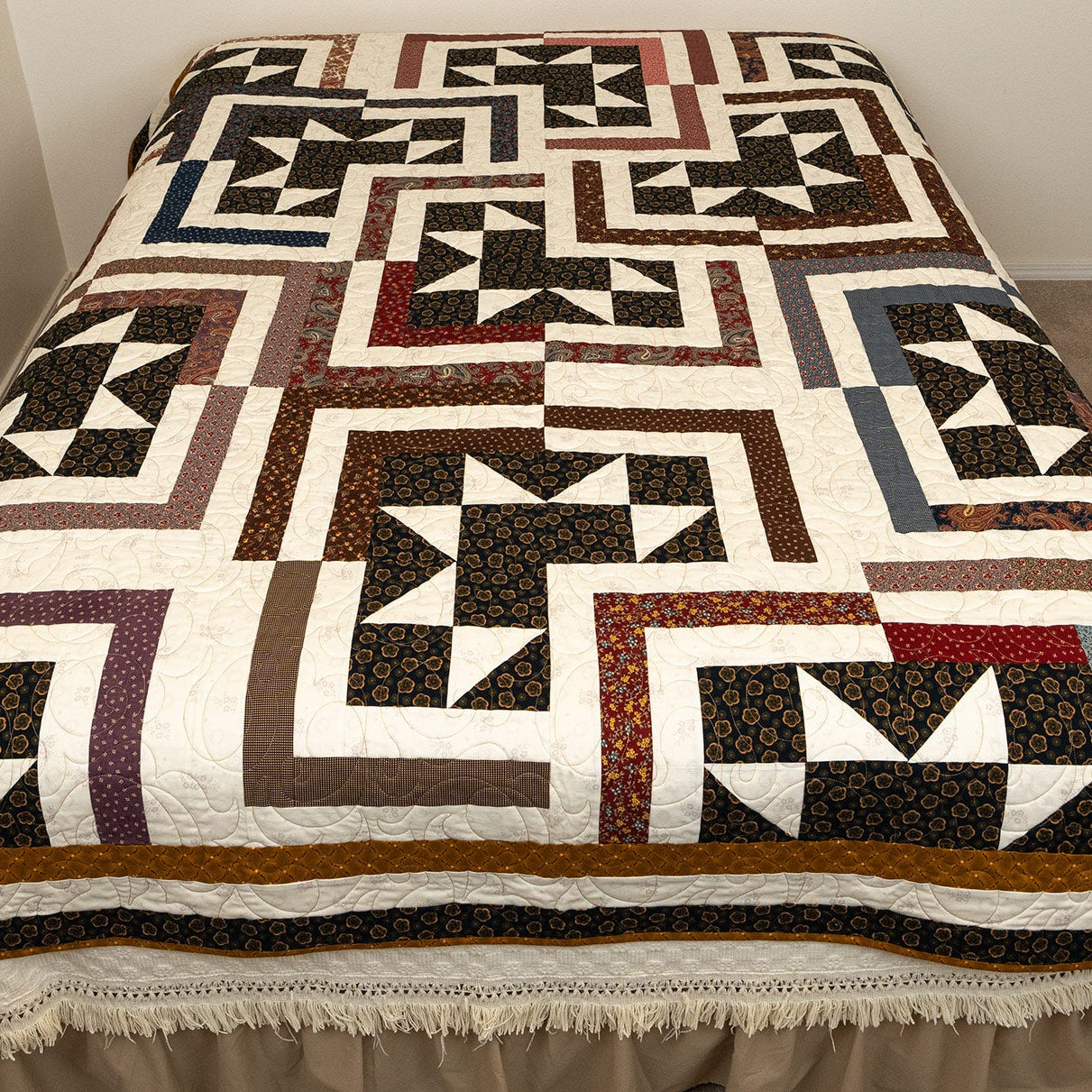A large quilted bedspread with a geometric block pattern in black, white, brown, and muted red, styled on a bed with a beige skirt in a bedroom setting.