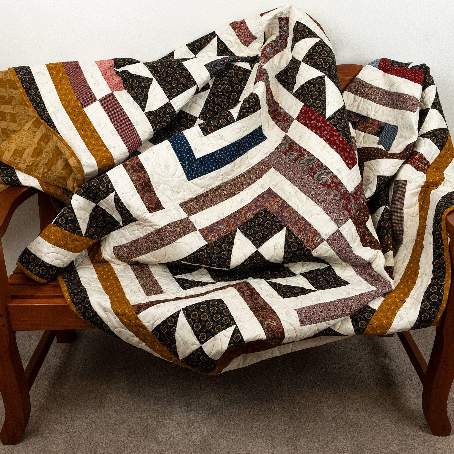 A large geometric patchwork quilt draped over a wooden armchair in a home setting, featuring a mix of brown, cream, black, and rust-orange patterns.
