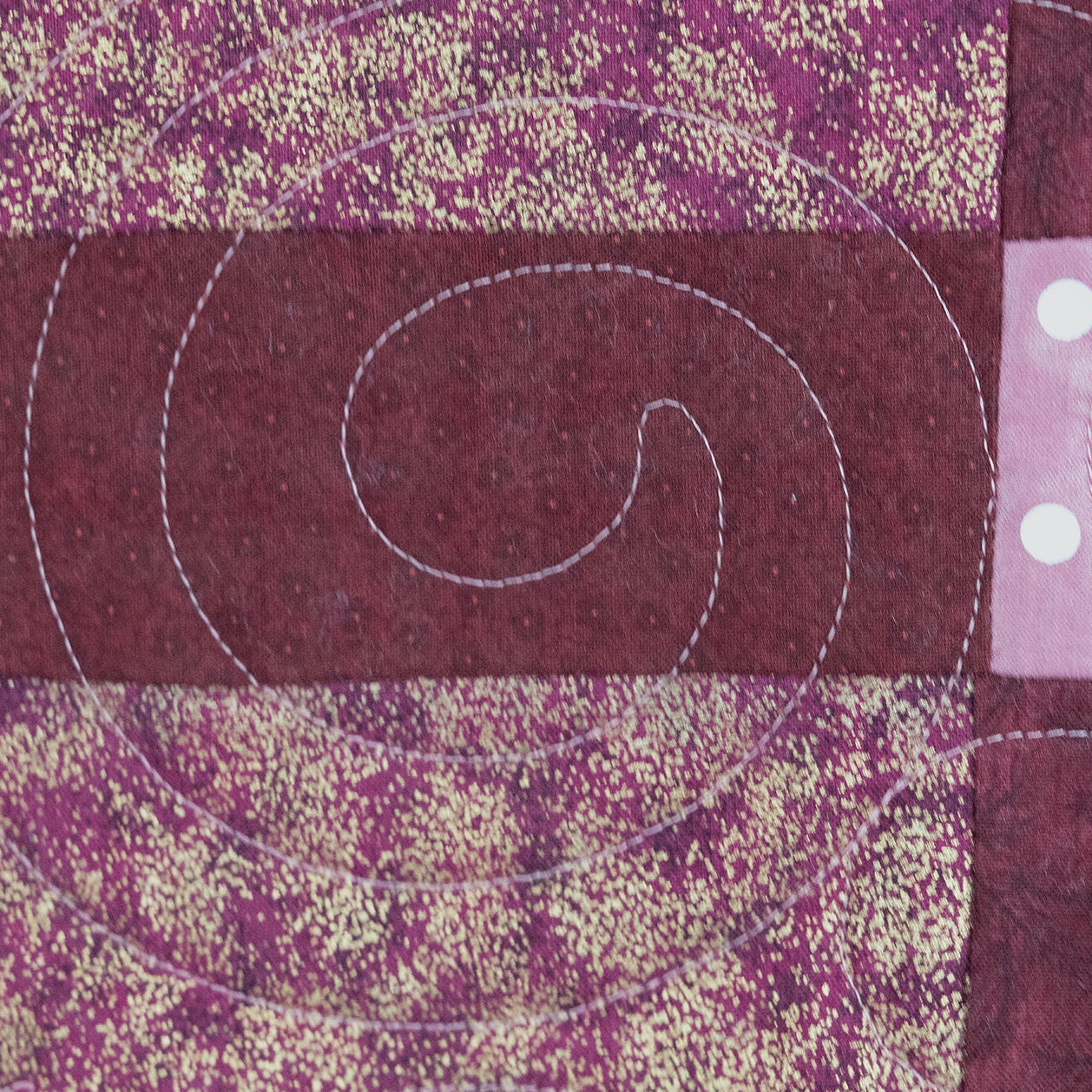 A close-up of a quilted fabric featuring a deep burgundy central panel with white curved quilting lines, flanked by purple and pink patterned sections with white polka dots.