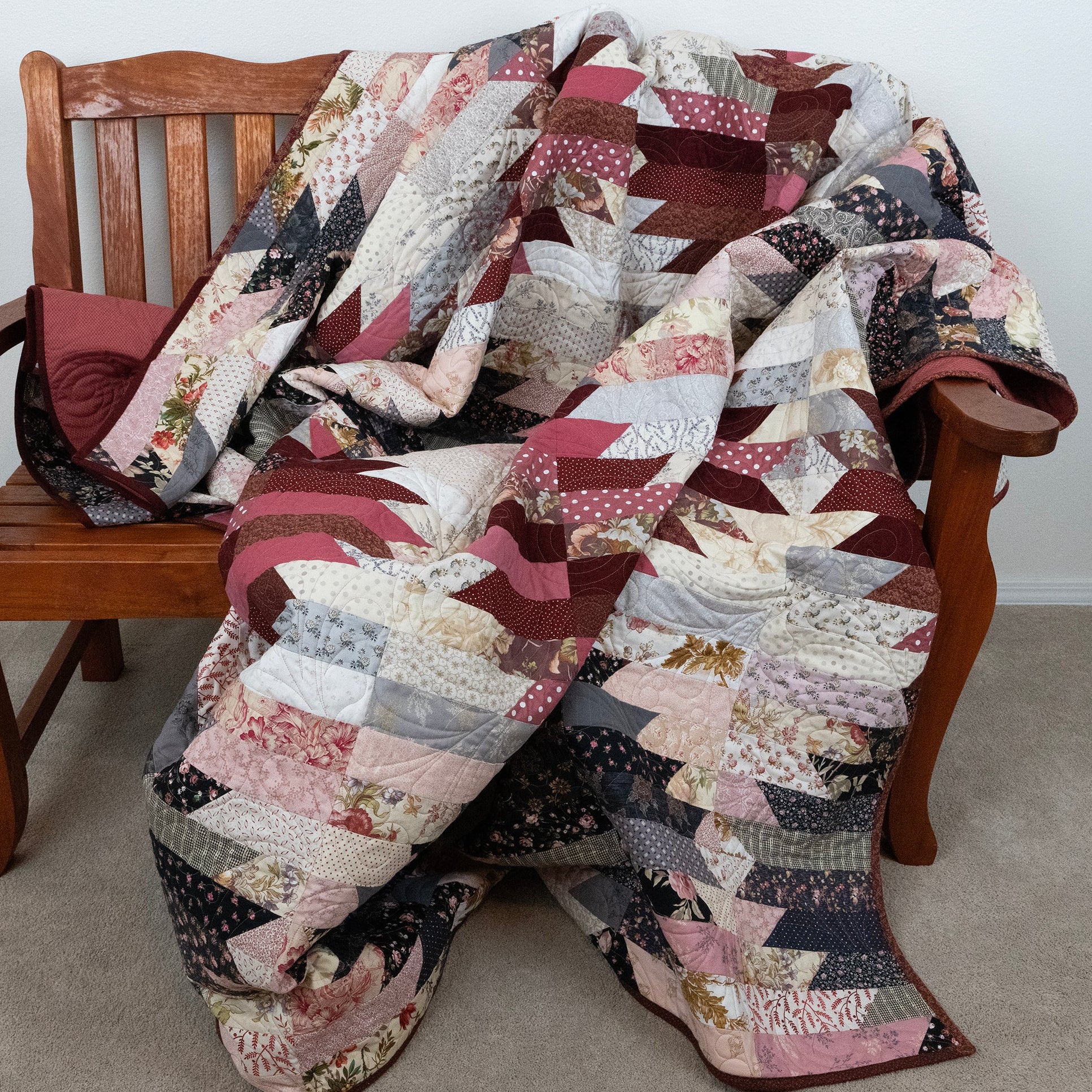A patchwork quilt draped over a wooden bench in an indoor room setting. The quilt features a mix of geometric and floral patchwork designs in dusty pink, burgundy, beige, gray, and black.