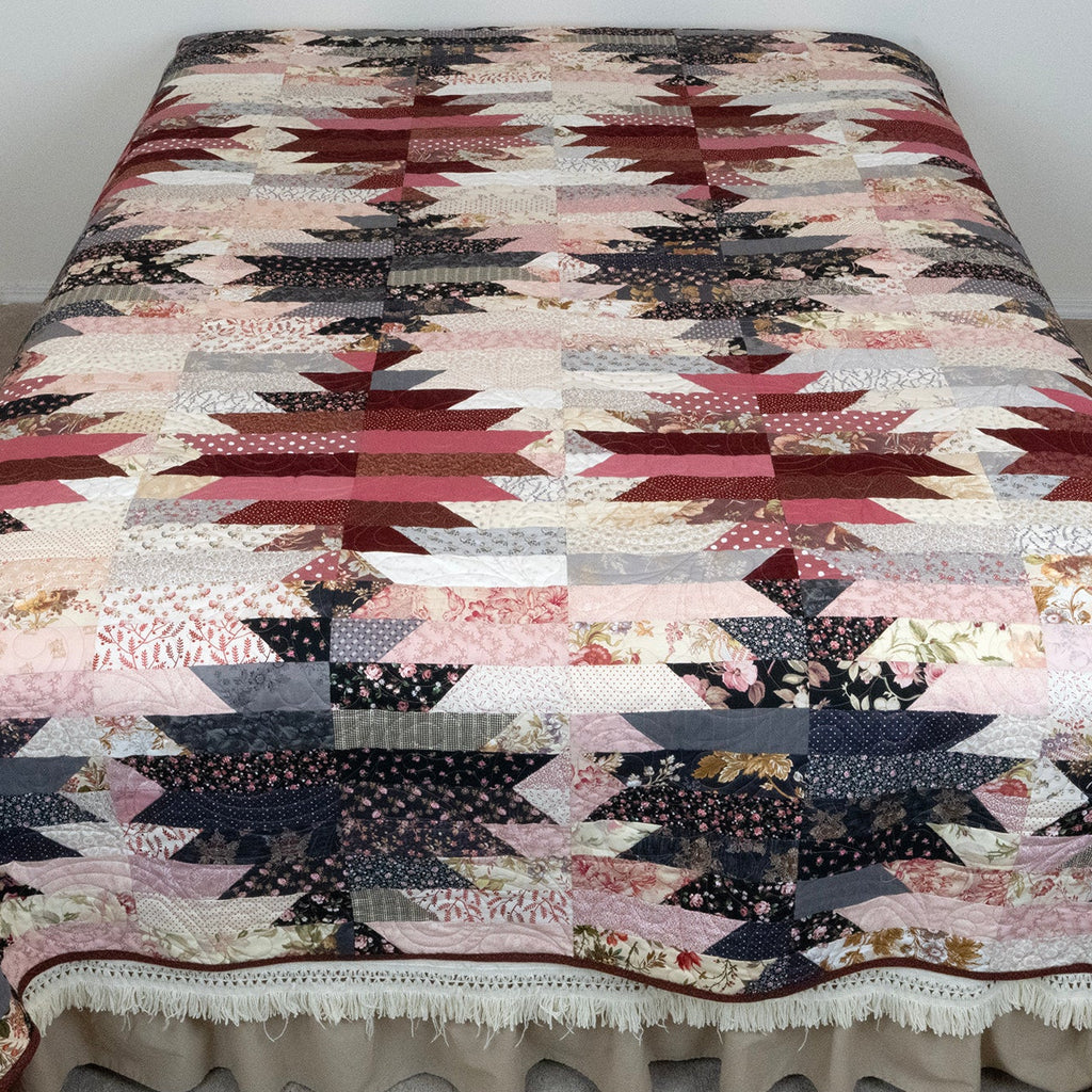 A patchwork quilt with a geometric starburst pattern in muted red, pink, beige, and navy tones, draped over a bed with a white ruffled bed skirt in a bedroom setting.