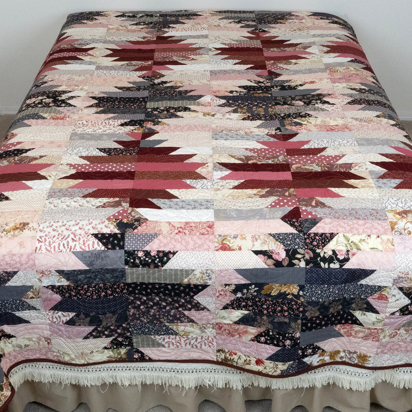 A patchwork quilt with a geometric starburst pattern in muted red, pink, beige, and navy tones, draped over a bed with a white ruffled bed skirt in a bedroom setting.