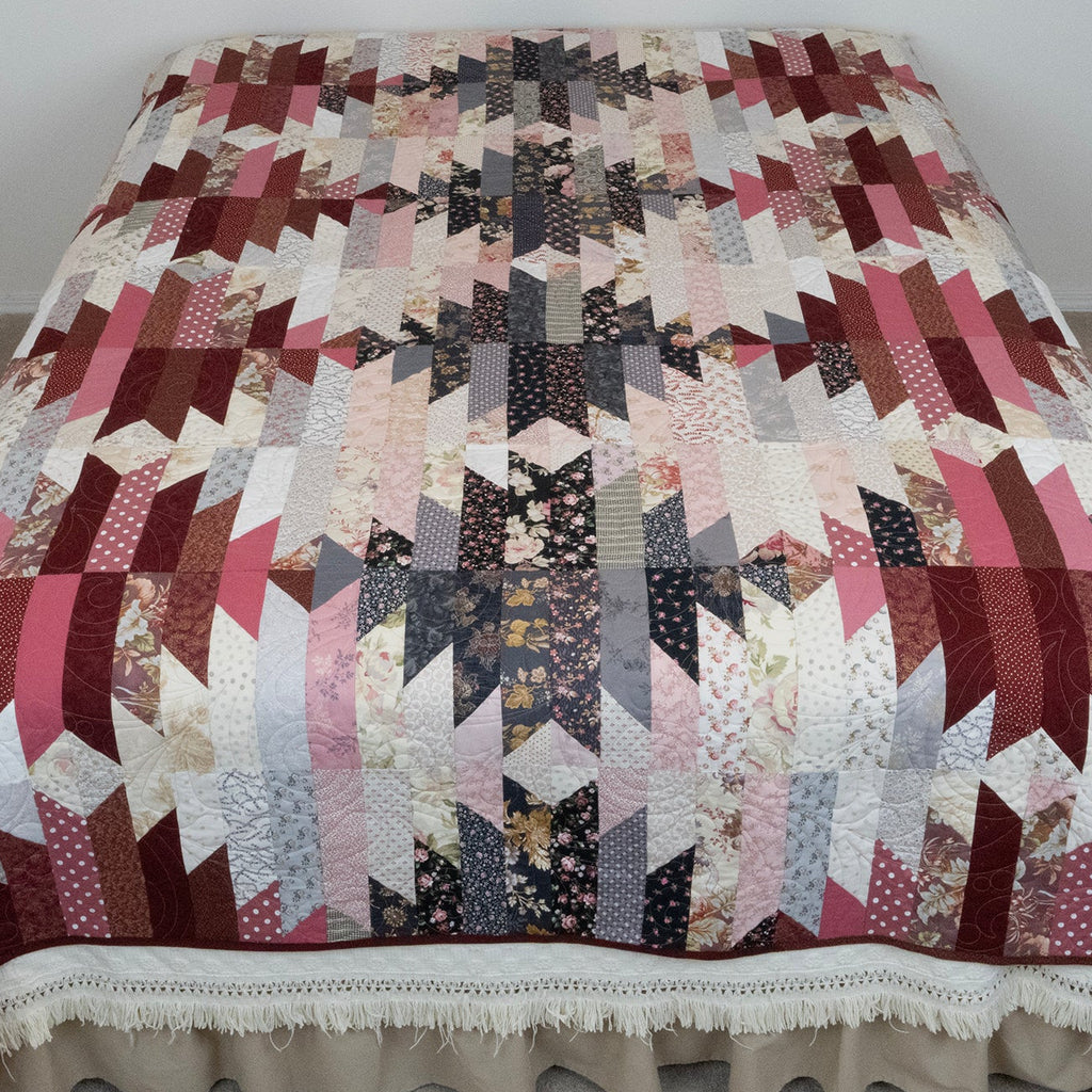 A quilted bedspread with a geometric patchwork design in shades of pink, burgundy, beige, and black, featuring a ruffled cream bed skirt, shown on a bed in a bedroom.
