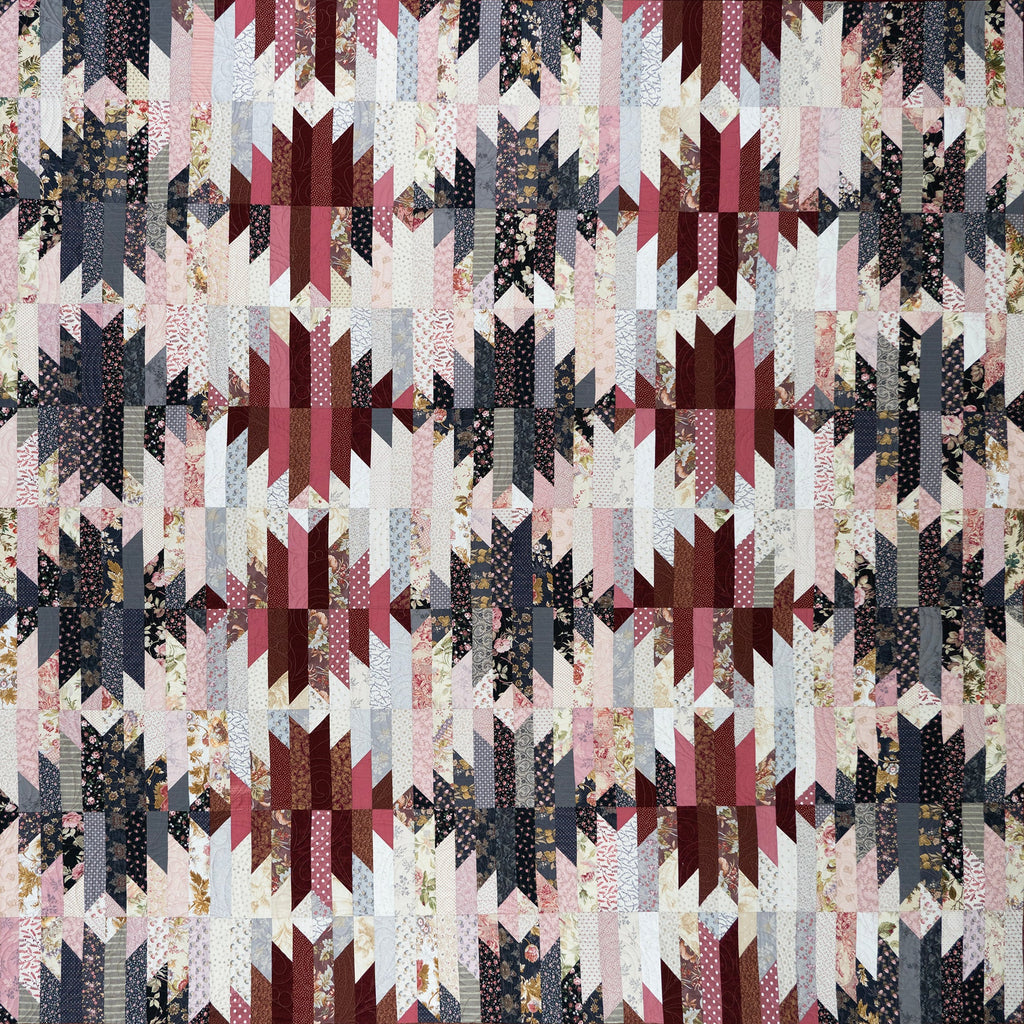 A square quilted textile with a repeating geometric patchwork pattern in muted pink, burgundy, charcoal gray, cream, and small floral print fabrics