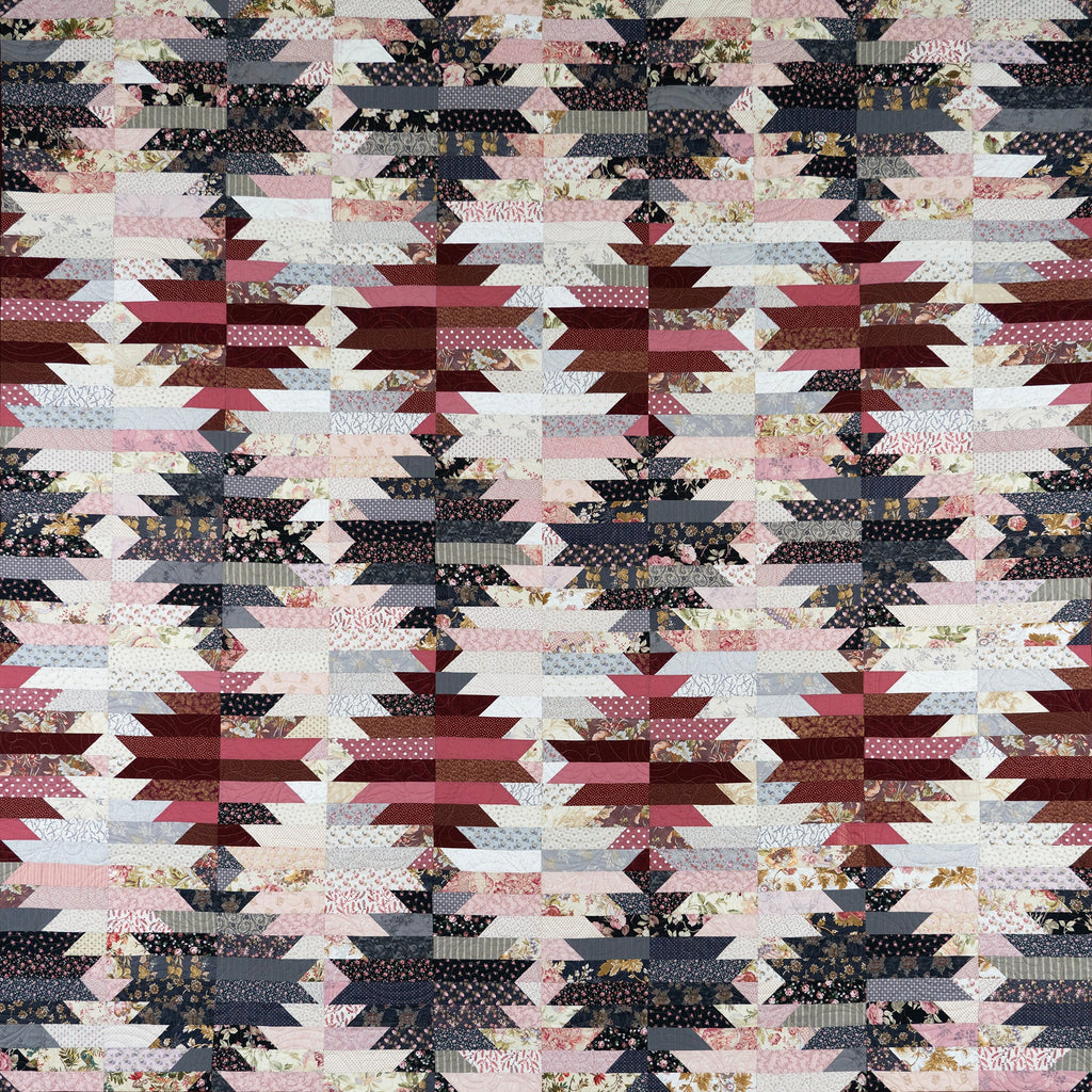 A square quilted textile featuring a repeating geometric star and diamond pattern in dusty pink, burgundy, charcoal gray, cream, and small floral print fabrics, finished with a dark border.