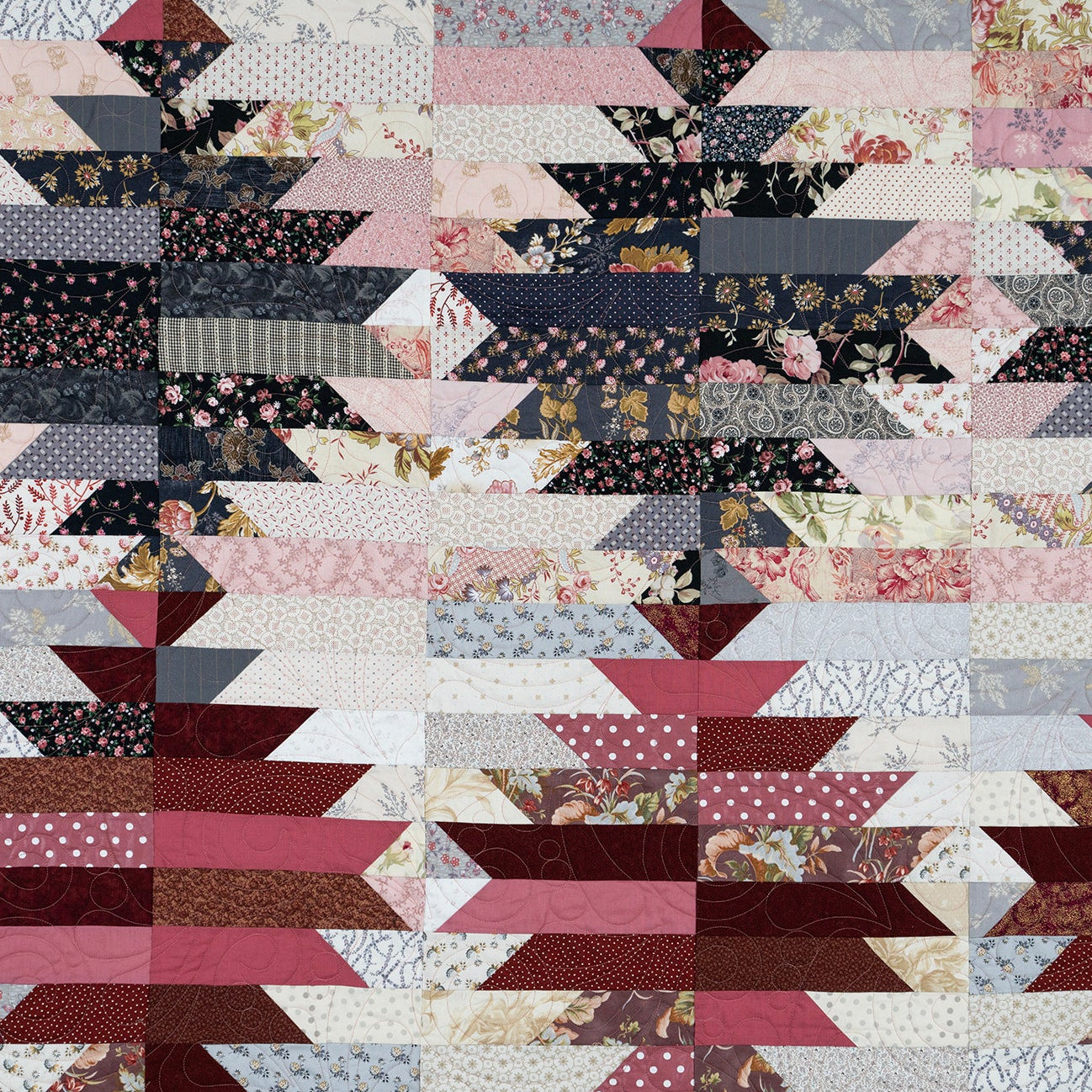 A close-up of a quilted fabric pattern featuring repeating geometric blocks with floral and striped textiles in muted pink, burgundy, navy, beige, and cream tones.