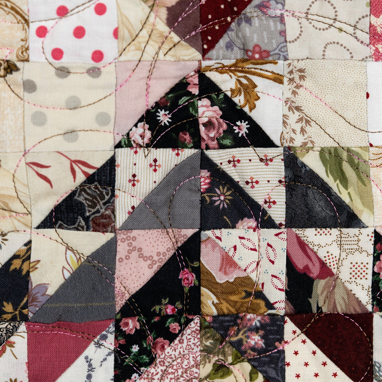 A close-up of a patchwork quilt featuring triangular fabric blocks with floral, polka dot, gingham, and paisley patterns in muted tones of pink, beige, black, and brown.