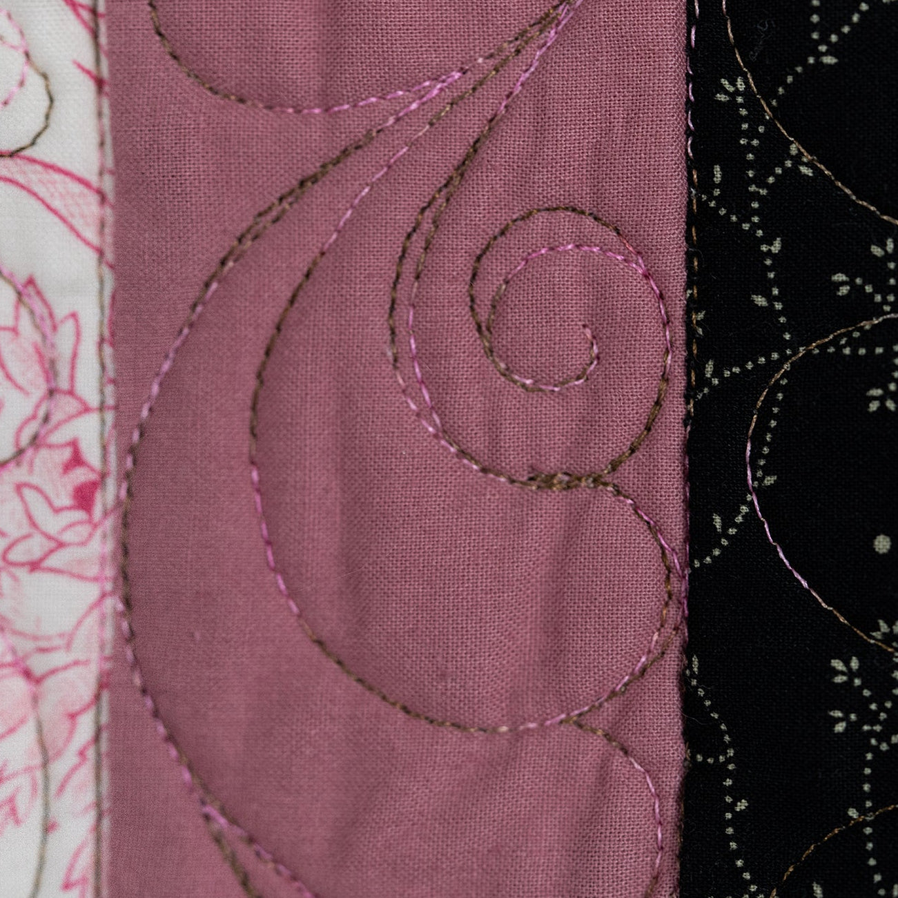 Close-up of a quilted fabric panel with three vertical sections: white with pink floral embroidery, dusty pink with swirling quilting, and black with white floral embroidery.