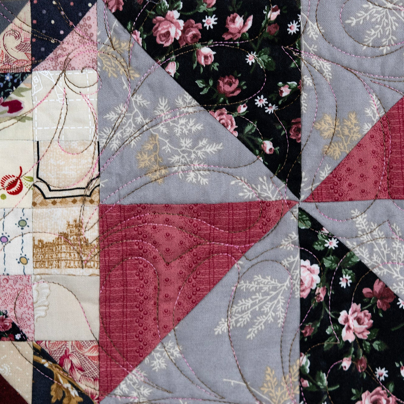 A close-up of a quilted patchwork textile with triangular fabric sections in dusty rose, light gray, black floral print, cream, and small gold accents, arranged in a geometric star-like pattern.