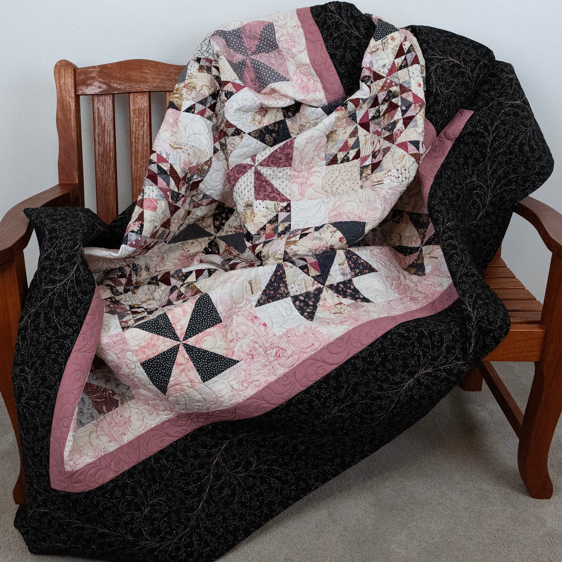 A patchwork quilt draped over a wooden chair in a home setting. The quilt features a complex geometric pattern with pink, black, and cream sections, including star and triangle motifs, bordered by a dusty pink stripe and a solid black border.