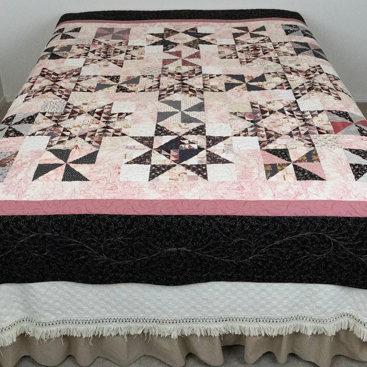 A quilted bedspread with a geometric starburst pattern in black, pink, and cream, draped over a bed with a white ruffled bed skirt in a simple room setting.