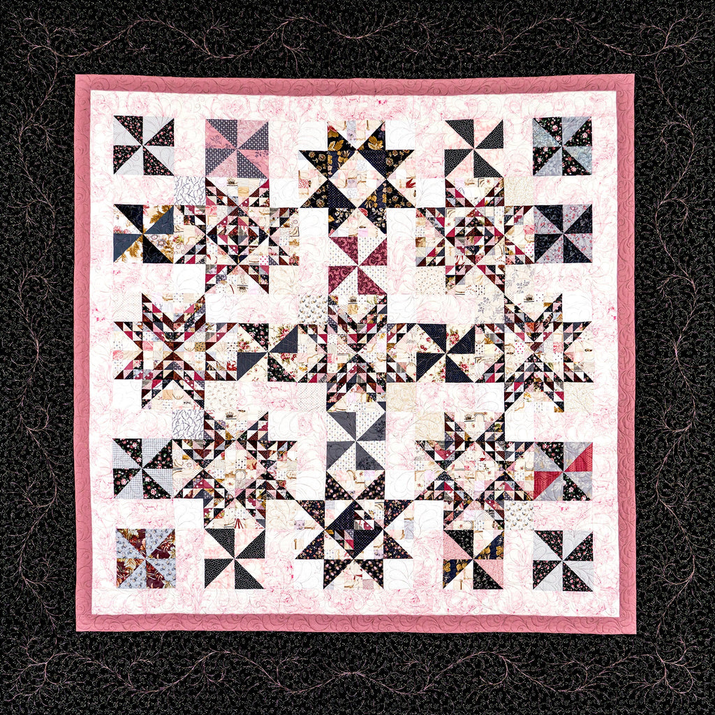 A square quilted textile with a pink border and a repeating geometric star-and-square block pattern in black, white, pink, and some multicolor fabric accents, shown flat against a dark patterned background.