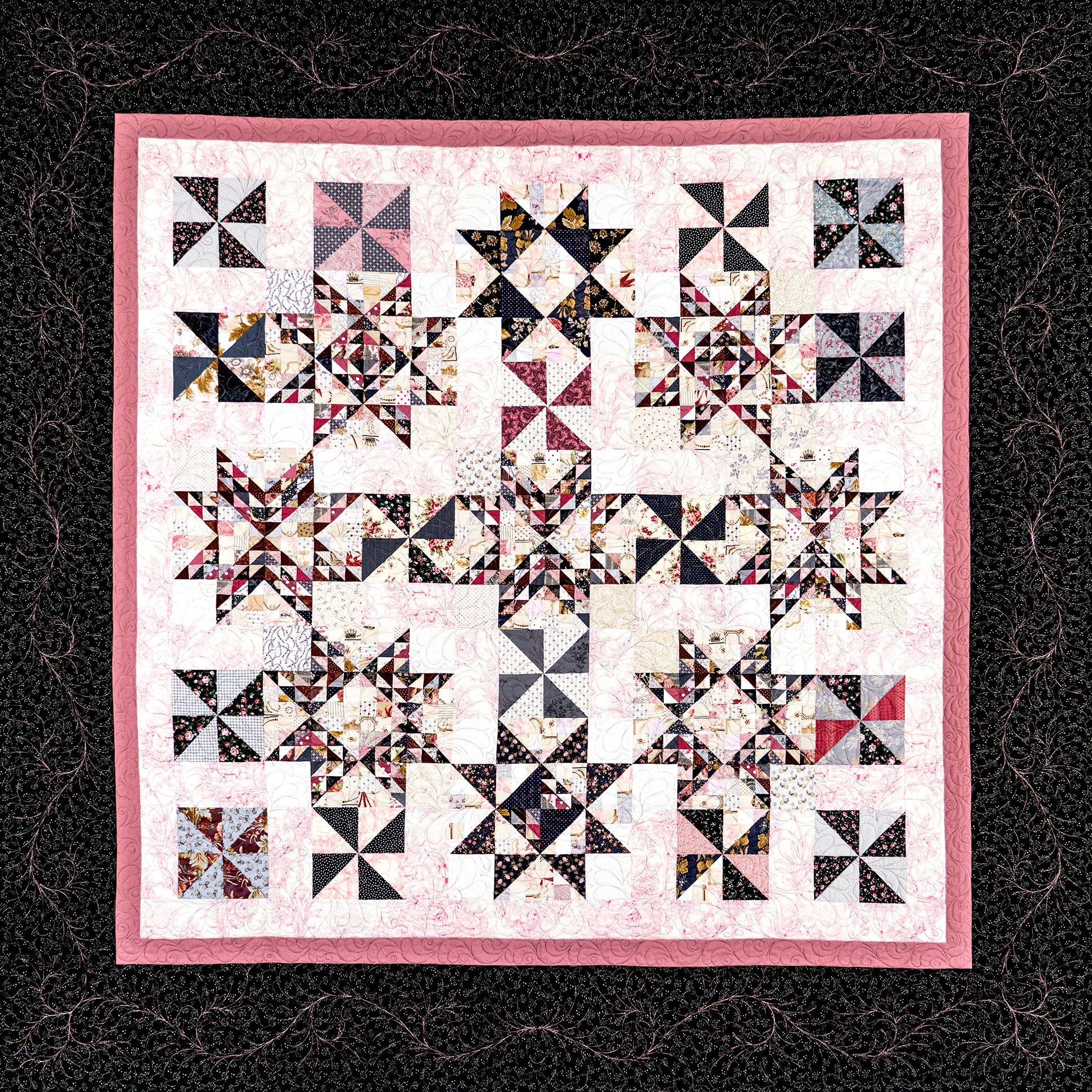 A square quilted textile with a pink border and a repeating geometric star-and-square block pattern in black, white, pink, and some multicolor fabric accents, shown flat against a dark patterned background.