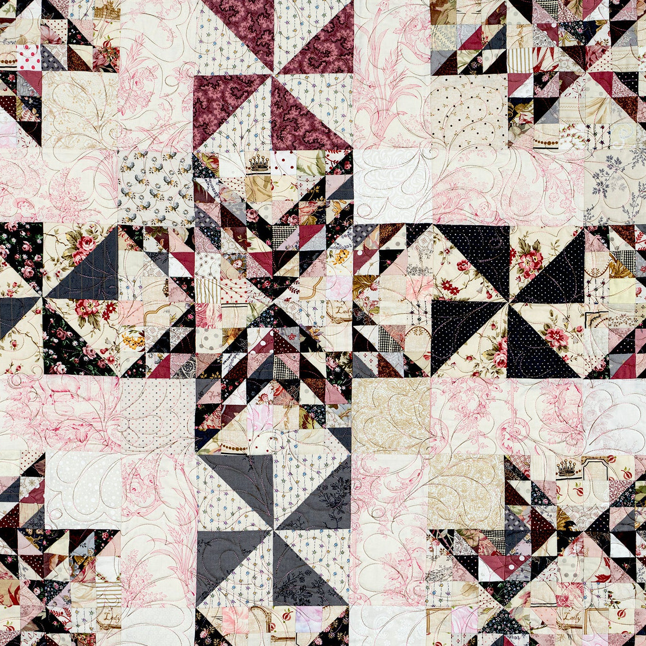 A square quilted textile with a patchwork design of geometric and pinwheel-style blocks in black, white, blush pink, and beige floral fabrics.