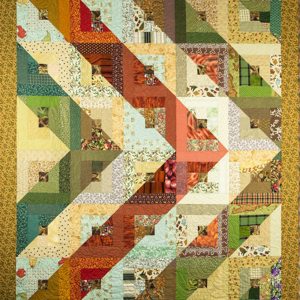A square quilted patchwork blanket featuring a geometric zigzag pattern made of various fabric panels in muted green, beige, orange, red, and brown tones, finished with a textured gold border.