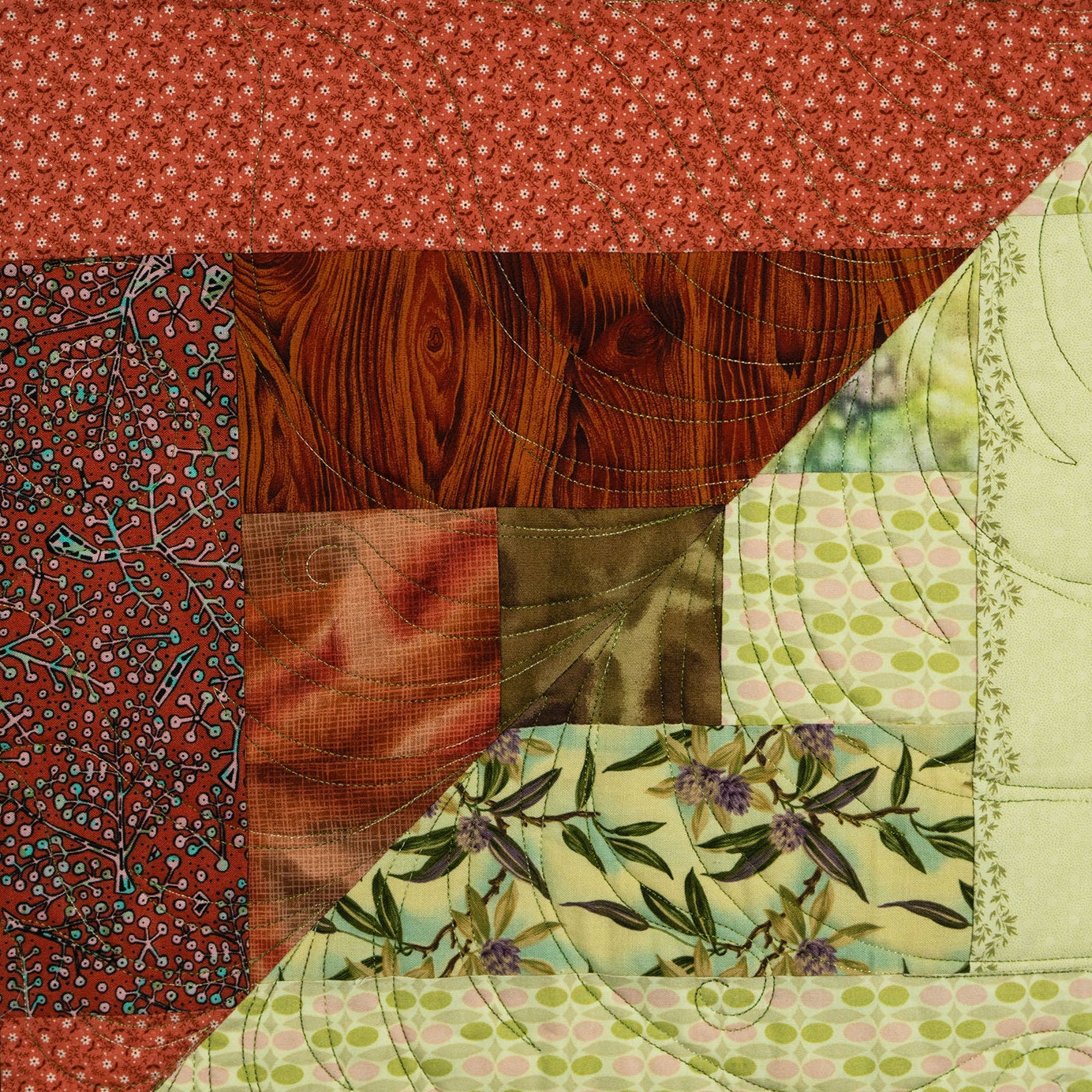 A close-up of a patchwork quilt featuring multiple fabric patterns, including red apple print, red floral fabric, brown wood grain, green and pink polka dots, and olive green leafy vines.