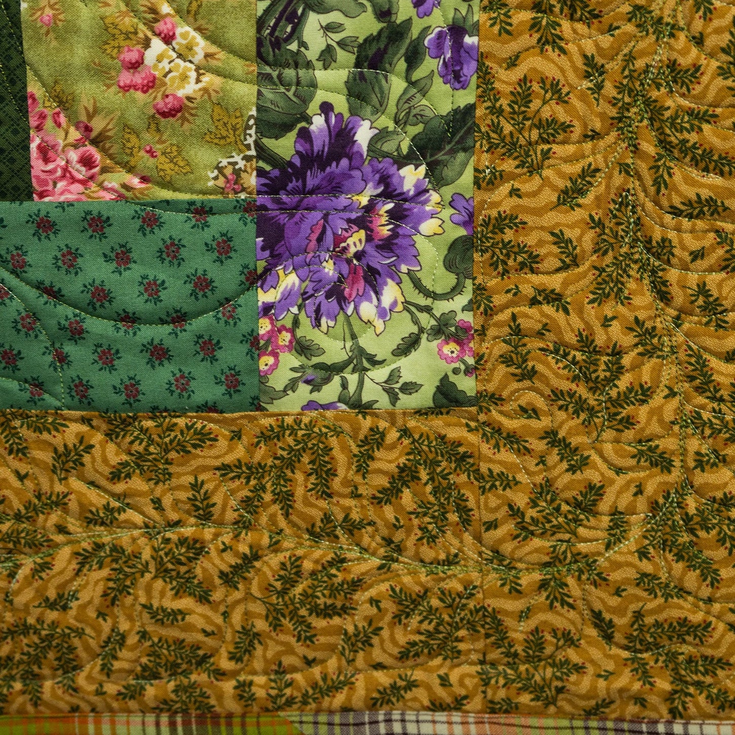 A close-up of a quilted fabric panel with a patchwork design. The largest section is mustard yellow with a dark green leafy vine pattern, while the top-left corner features green floral and pink floral prints.