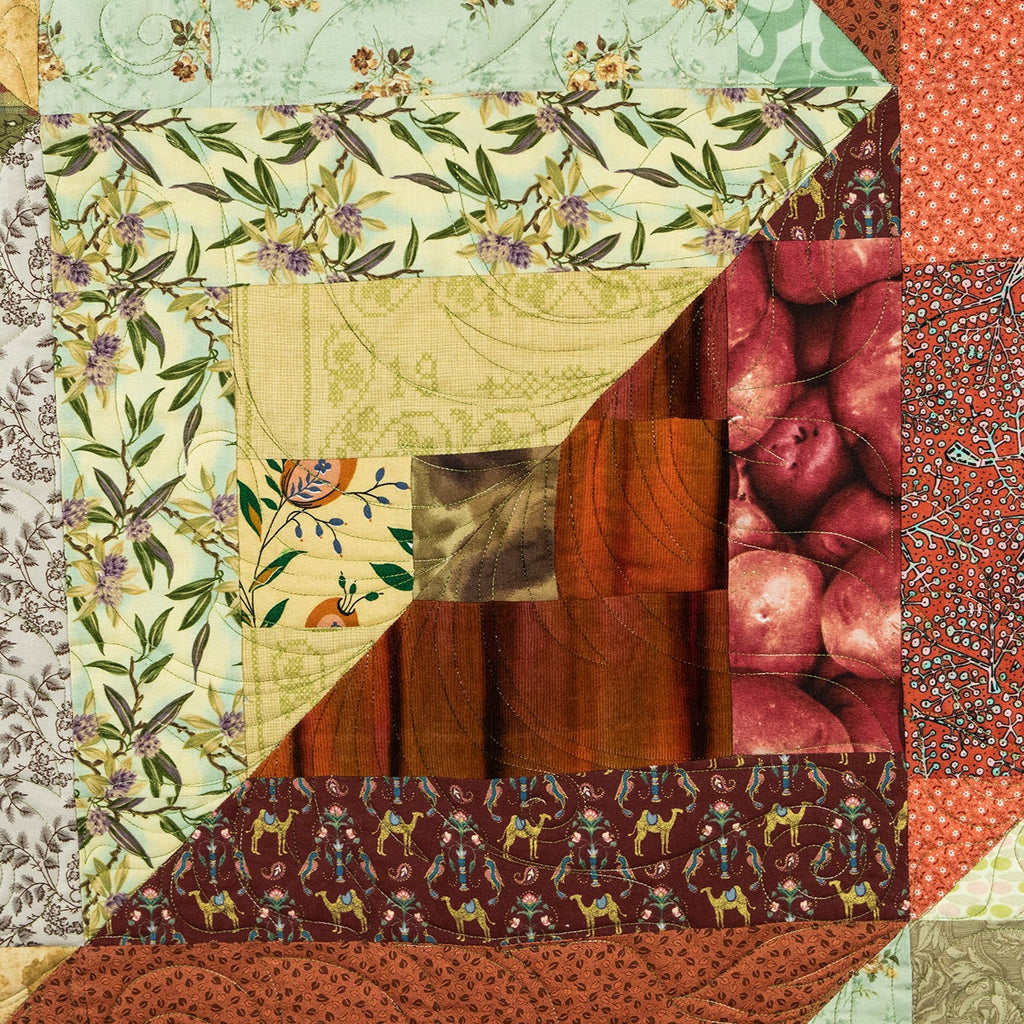 A close-up of a patchwork quilt featuring multiple fabric panels with different patterns, including floral prints, fruit motifs, text prints, and solid colors in red, green, beige, brown, and orange.