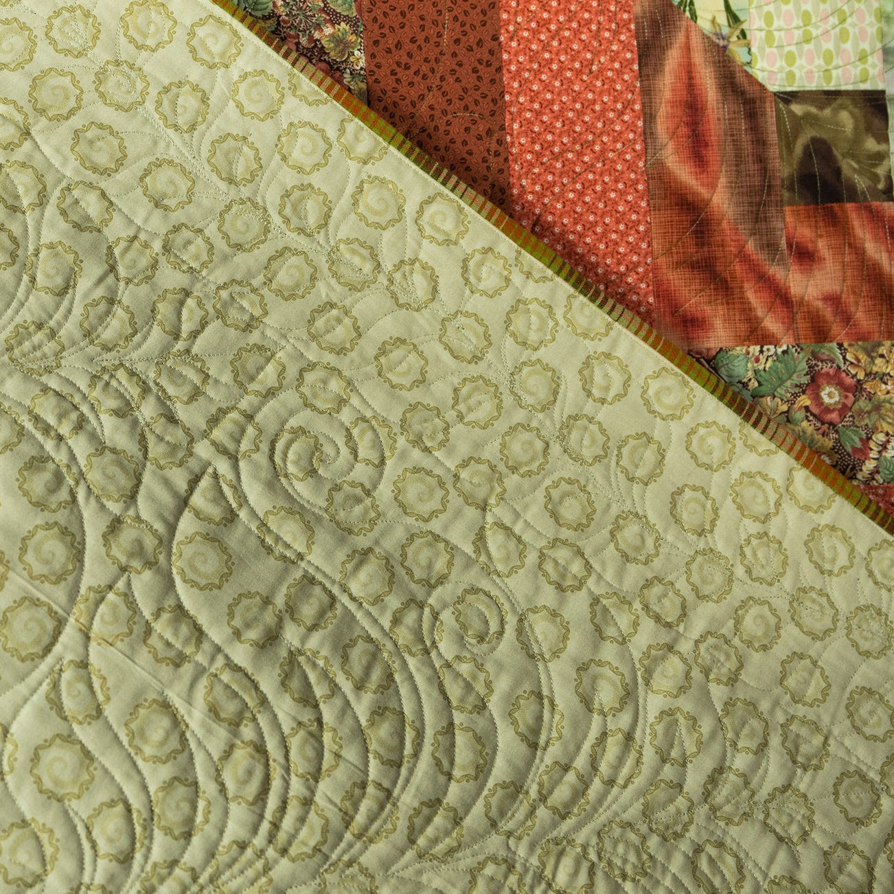 A close-up of a quilted fabric with swirling stitched patterns on a light cream background, with a diagonal section showing a red patterned fabric and a floral textile.