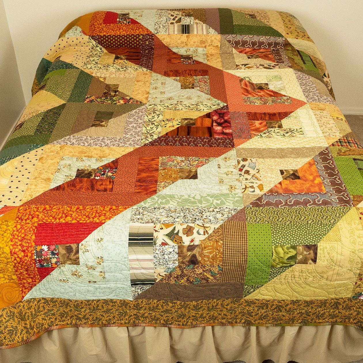 A multicolor geometric patchwork quilt draped over a bed with a beige ruffled bed skirt, set in a room with beige carpet and off-white walls.