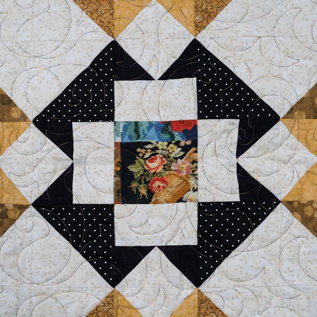A square quilted textile with a geometric patchwork design, featuring alternating diamond sections of black with white polka dots, white with light swirls, tan/gold patterned fabric, and a central floral print square.