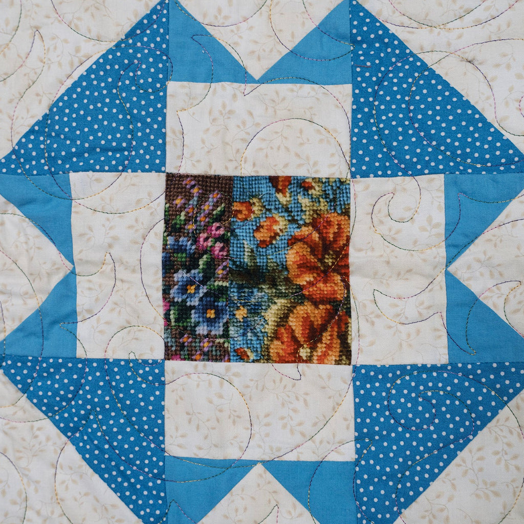 A close-up of a quilted textile with a geometric starburst pattern in shades of blue, white, and navy, featuring a central floral patch with orange, blue, and pink flowers.