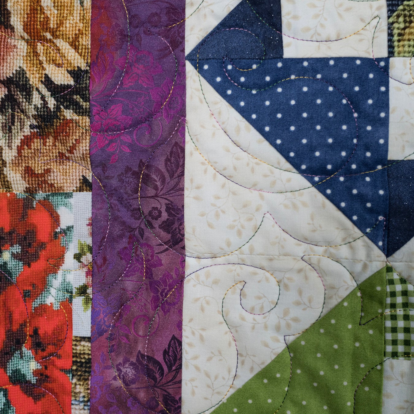 A close-up of a patchwork quilt showing multiple fabric panels with floral, paisley, polka dot, and gingham patterns in red, purple, navy, green, beige, and brown.