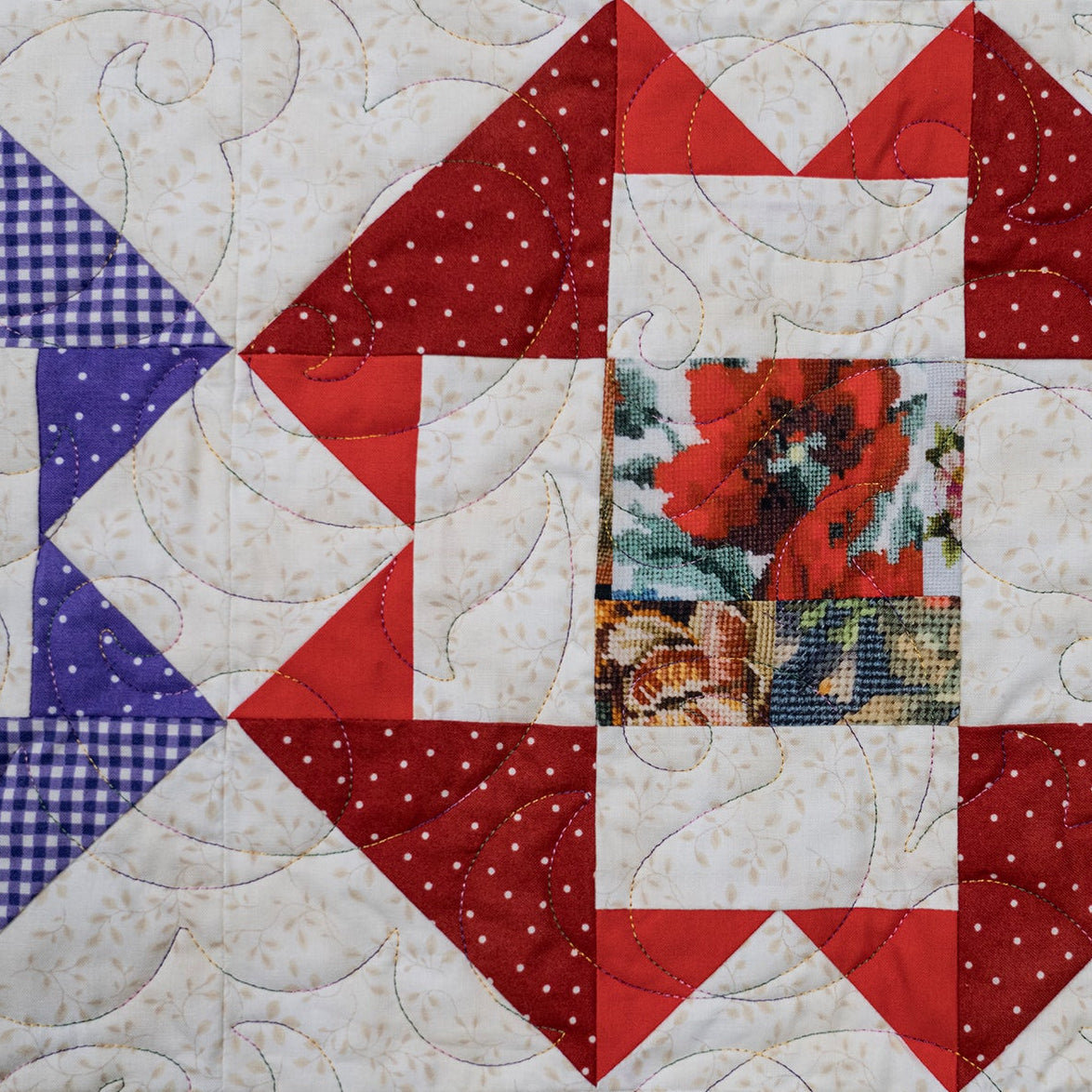 A square quilted patchwork textile with a geometric star-like pattern. The design includes red and white patterned triangles, a blue-and-white gingham section, a floral center patch all stitched together on a white background.