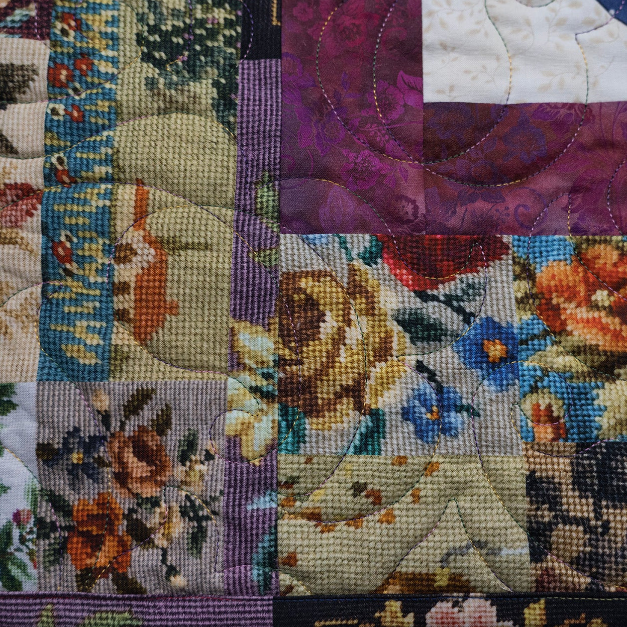 A close-up of a patchwork quilted textile featuring floral needlepoint-style panels in purple, orange, blue, green, and cream, with a dark border.