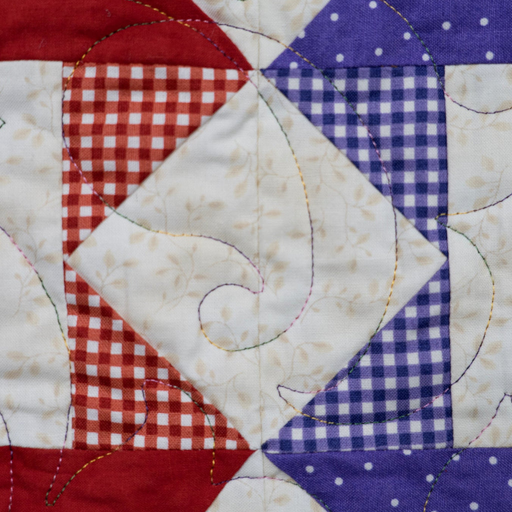 A close-up of a quilted fabric panel featuring a patriotic color-blocked design with red, white, and blue sections. The fabrics include solid red, red-and-white gingham, white with a subtle floral print, blue-and-white gingham, and blue with white polka dots.