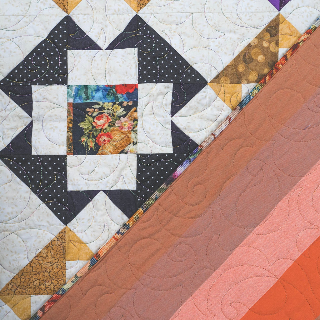A close-up of a quilted textile with a geometric patchwork design on the left and diagonal solid color bands on the right, including white, black, tan, floral multicolor, purple, brown, peach, and orange tones.
