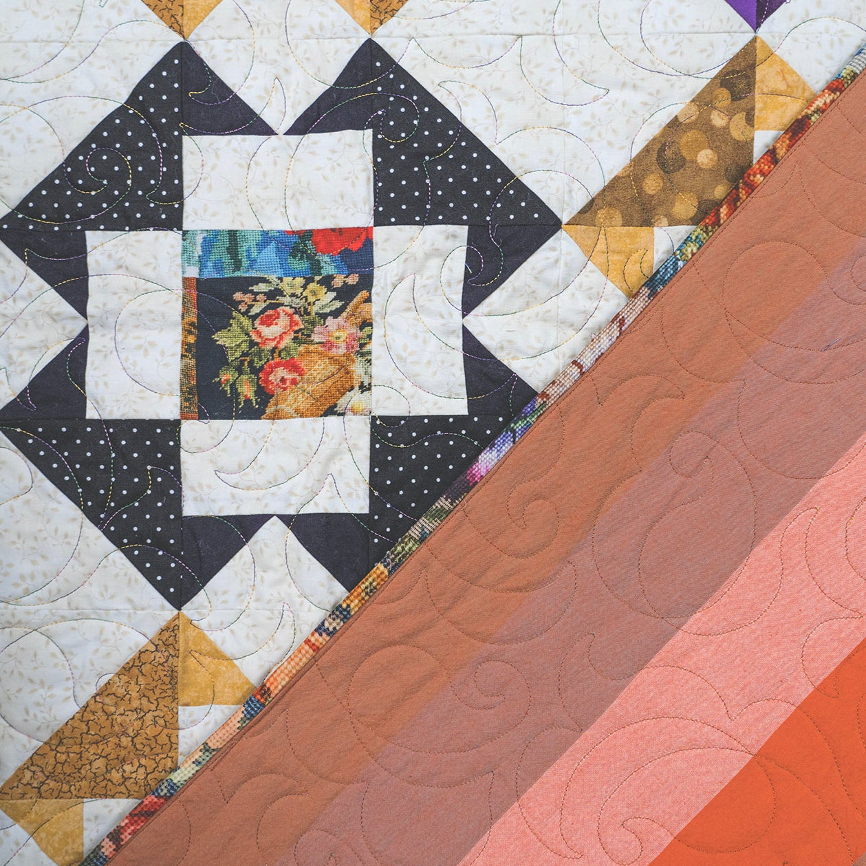 A close-up of a quilted textile with a geometric patchwork design on the left and diagonal solid color bands on the right, including white, black, tan, floral multicolor, purple, brown, peach, and orange tones.
