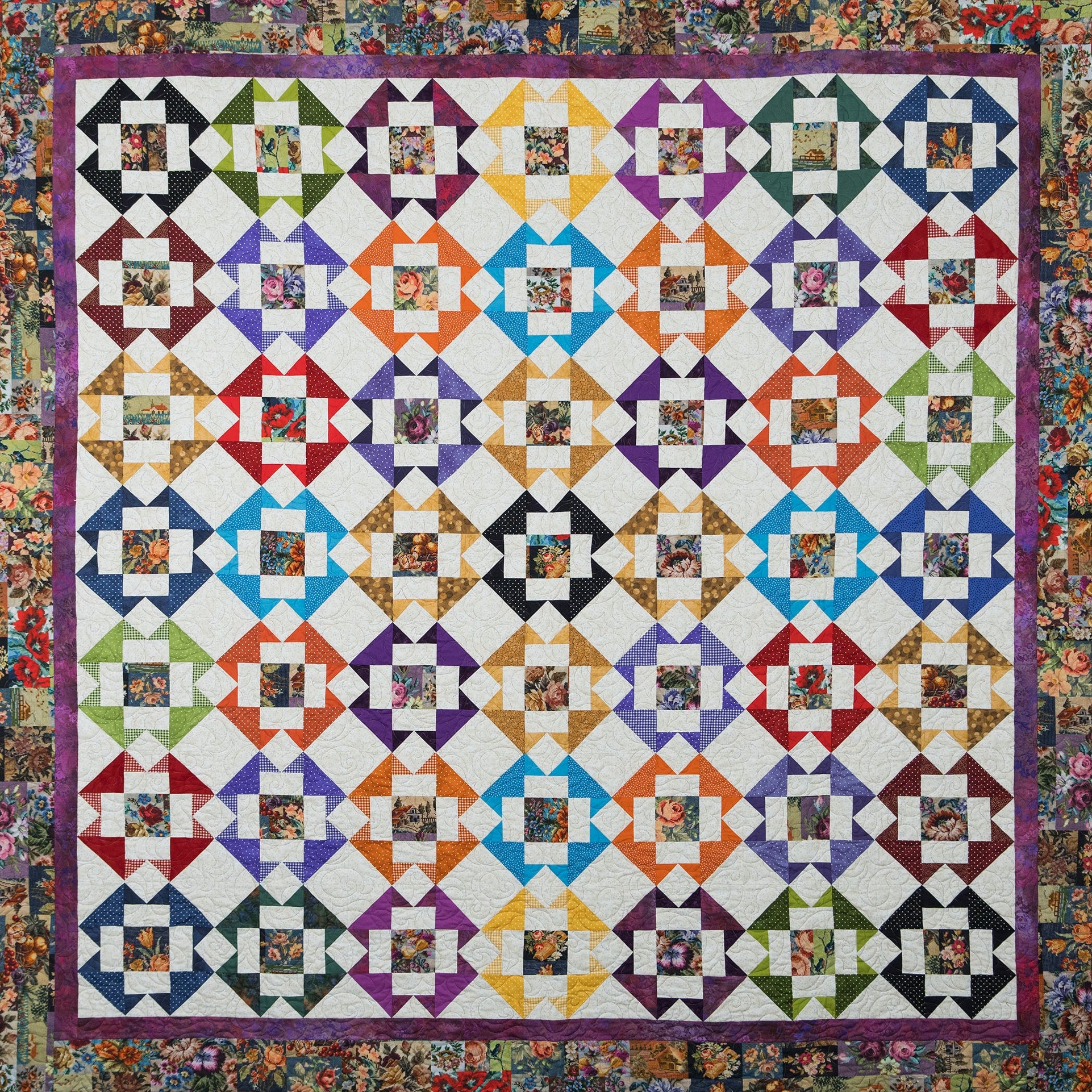 A square quilt with a colorful geometric block pattern arranged in rows on a white background, surrounded by a floral border. Each block contains small floral photographs, making the overall color identity highly multicolored.