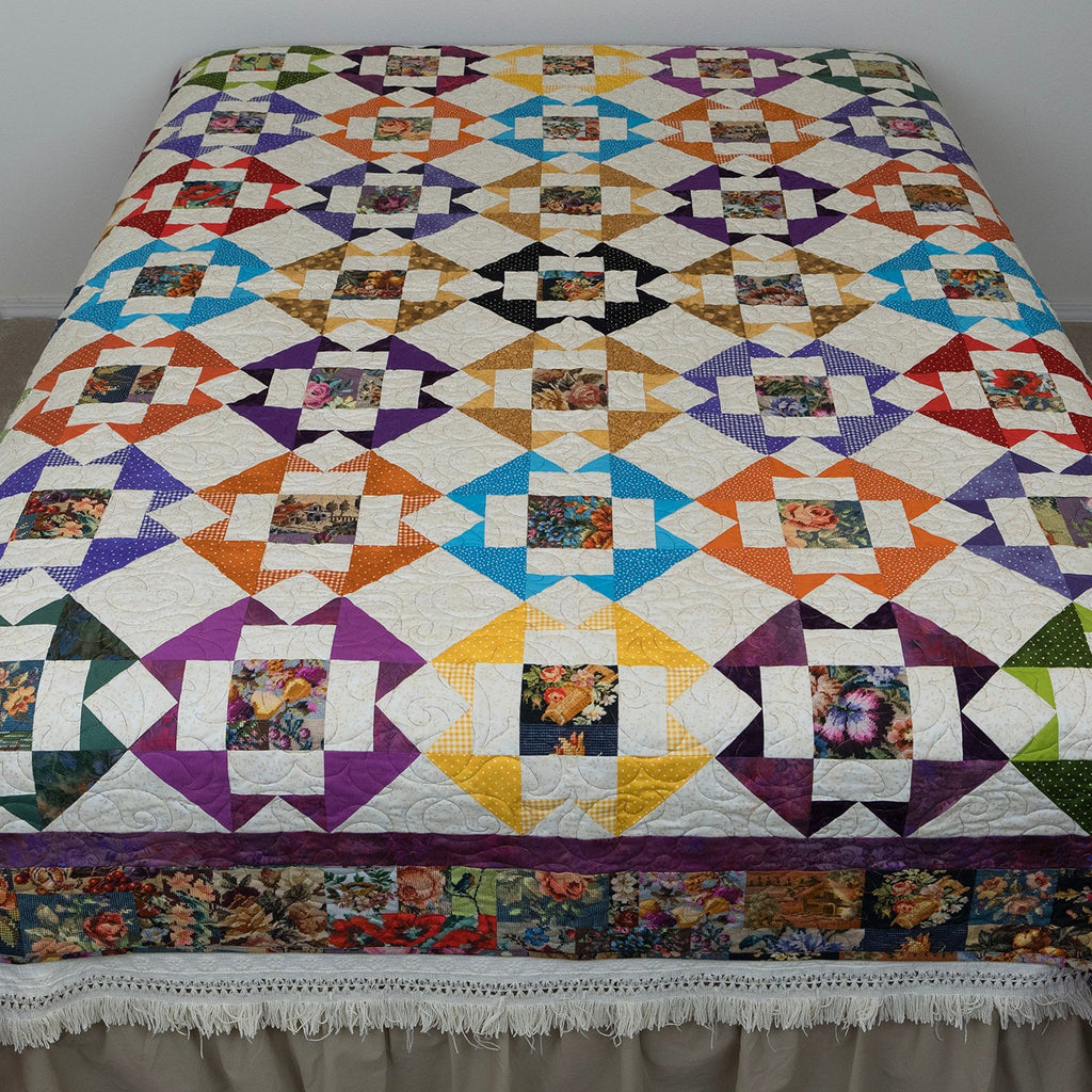A colorful quilted bedspread with a geometric patchwork pattern and a floral border, displayed on a bed in a simple room setting.