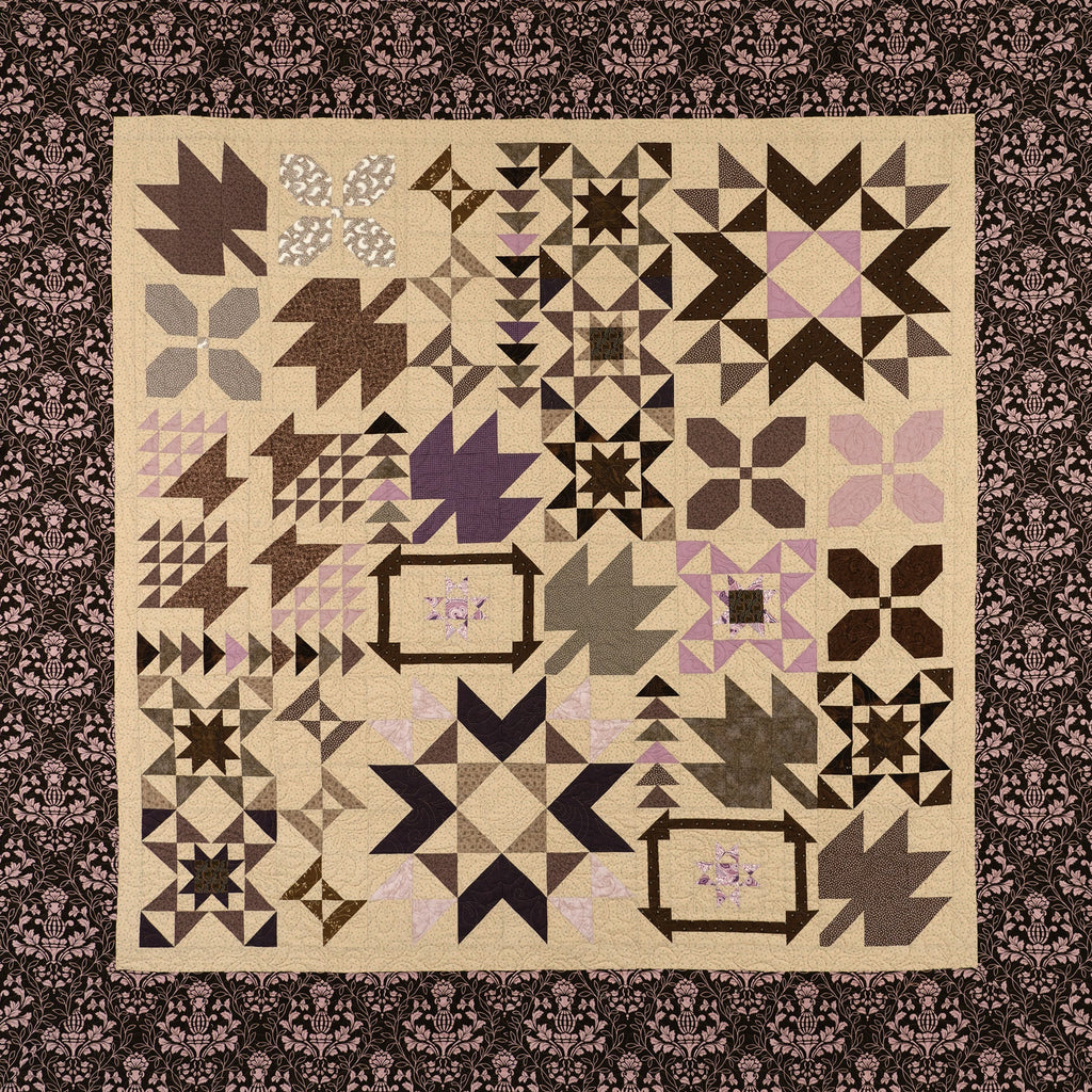 A square quilted textile with a patchwork arrangement of geometric and floral block patterns in beige, brown, black, gray, and soft pink, surrounded by a dark patterned border.