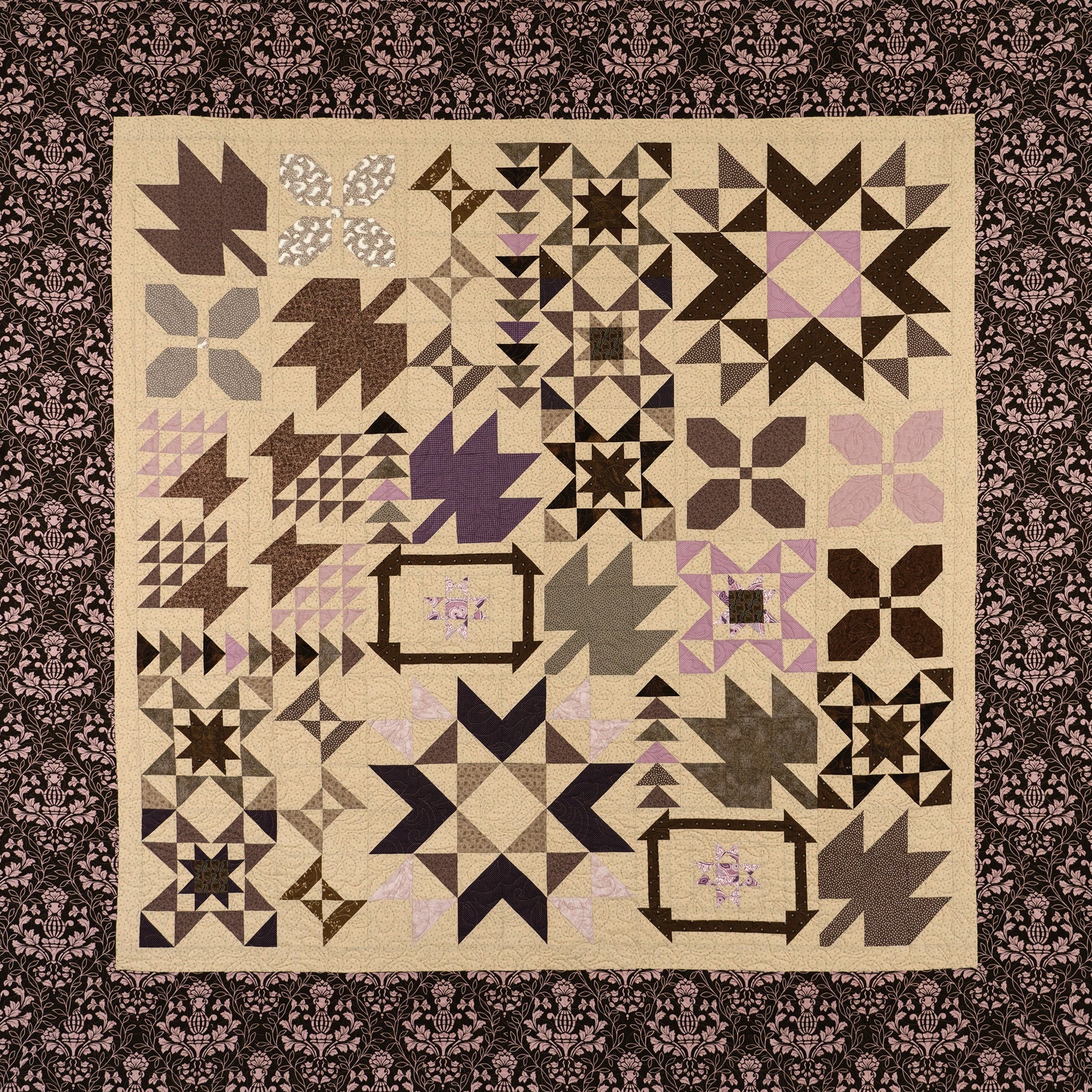 A square quilted textile with a patchwork arrangement of geometric and floral block patterns in beige, brown, black, gray, and soft pink, surrounded by a dark patterned border.