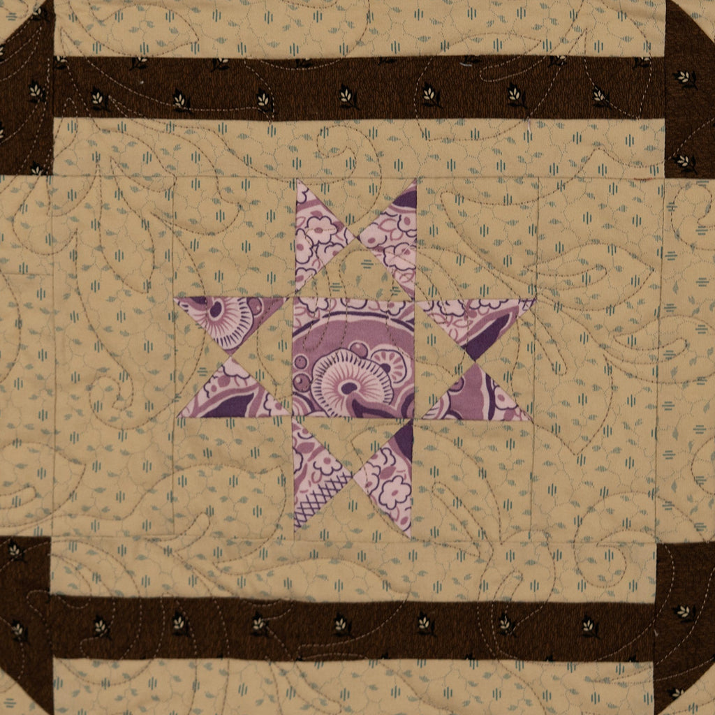 A square quilted textile with a geometric star design, featuring a beige patterned field, a purple paisley center star, and dark brown arrow-like borders with small black stitched details.