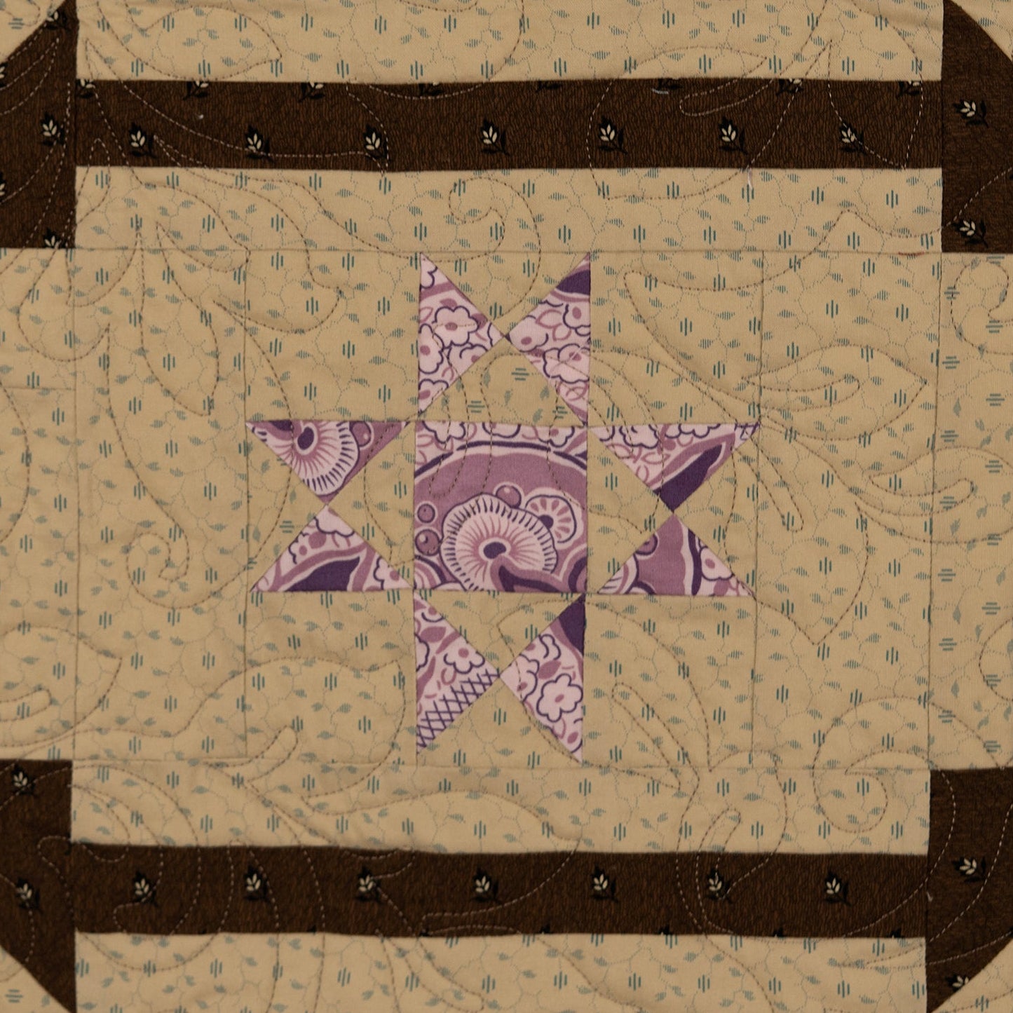 A square quilted textile with a geometric star design, featuring a beige patterned field, a purple paisley center star, and dark brown arrow-like borders with small black stitched details.