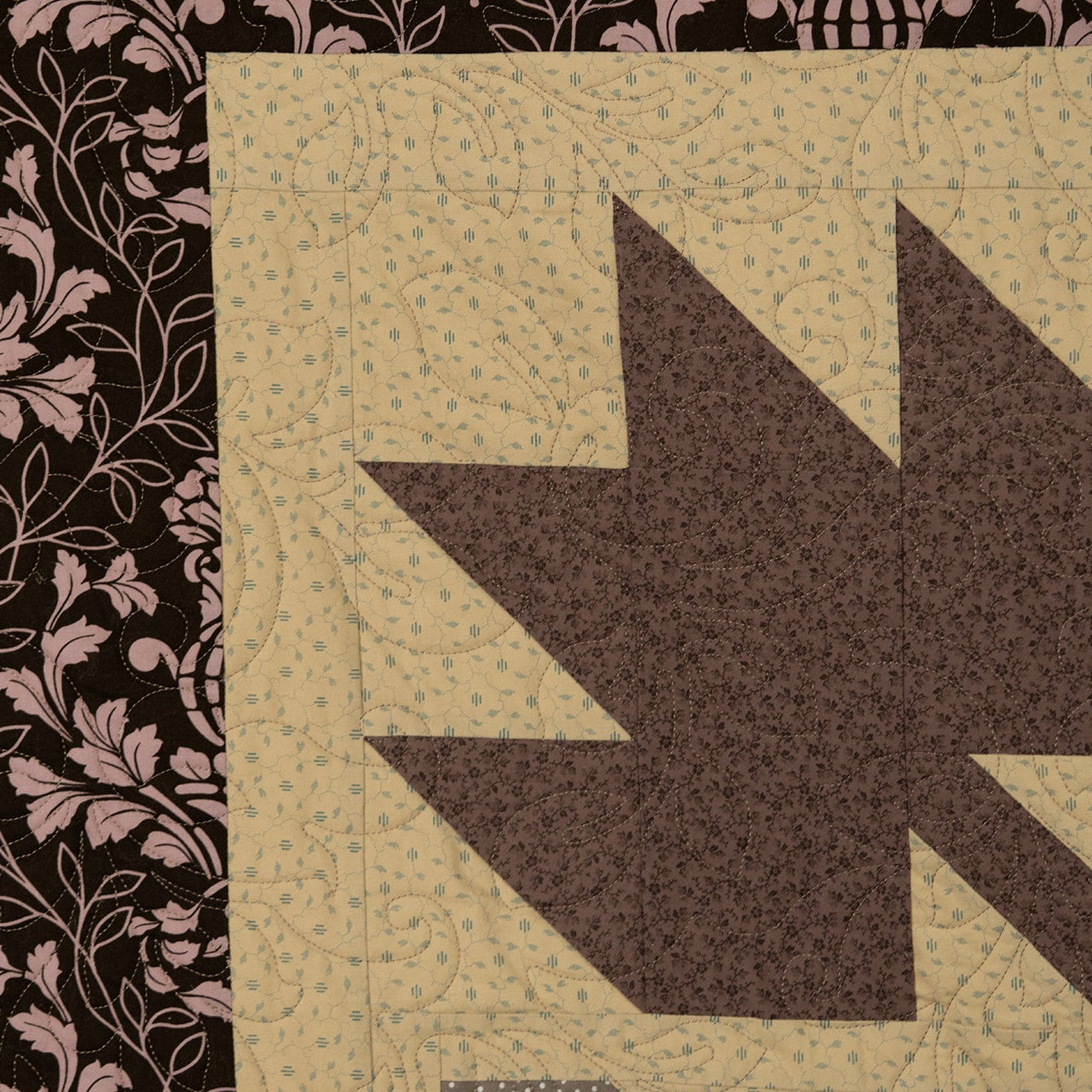 A close-up of a quilted fabric pattern featuring a large dark brown geometric maple leaf shape on a beige background, next to a black panel with a pink damask floral pattern.
