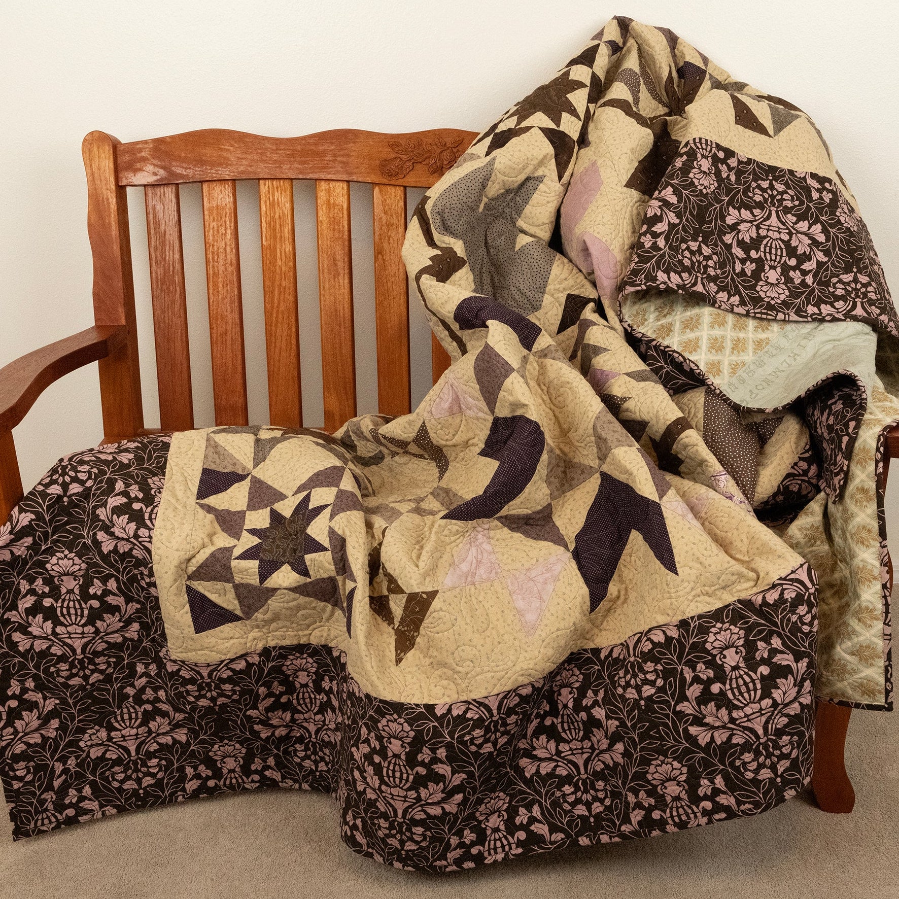 A patchwork quilt draped over a wooden bench in a home setting. The quilt features a mix of geometric star patterns and floral damask borders in beige, brown, and muted pink, with a light green patterned insert visible on the right side.