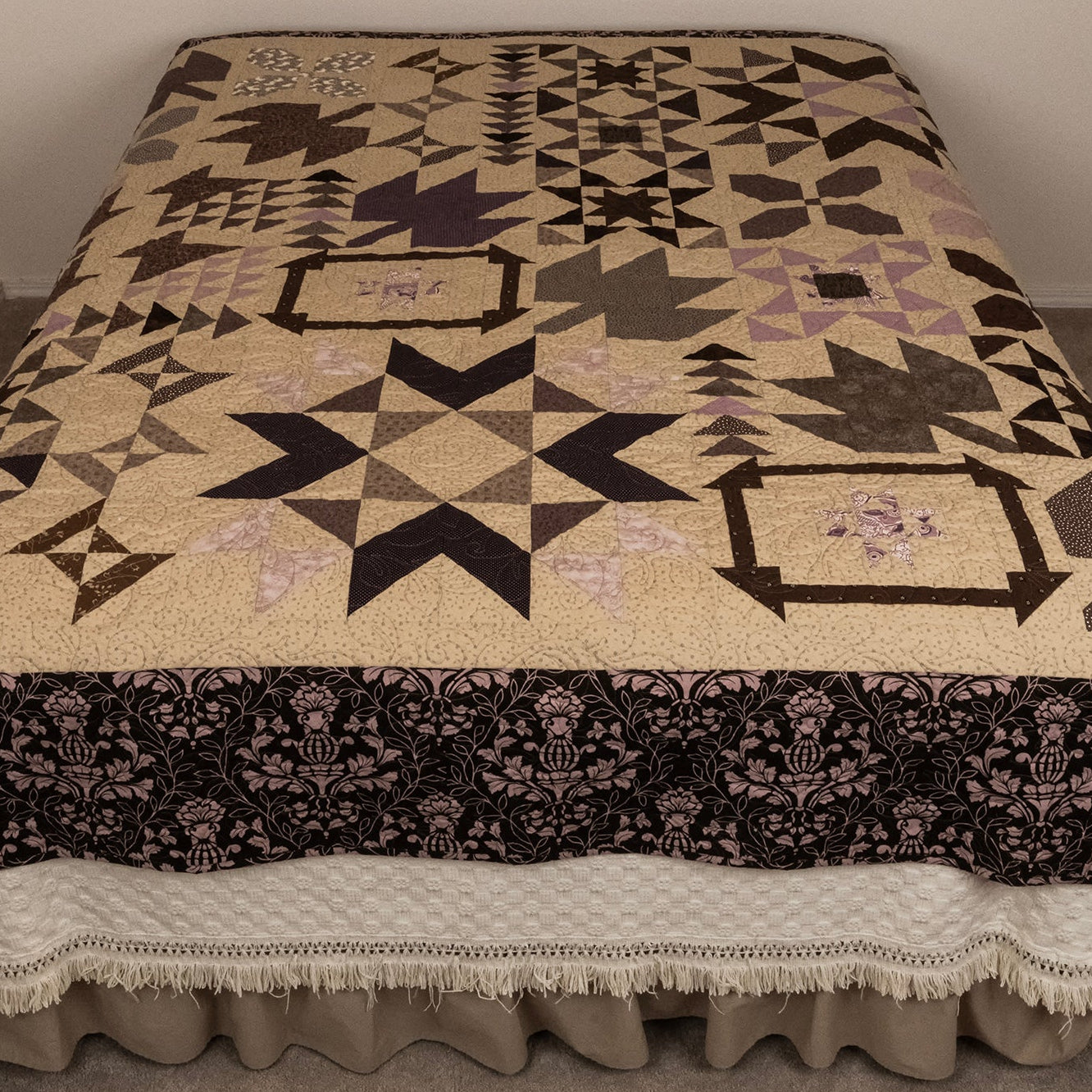 A large quilted bedspread with a geometric and floral pattern in beige, black, and muted purple, styled on a bed with a white ruffled bed skirt in a neutral room.