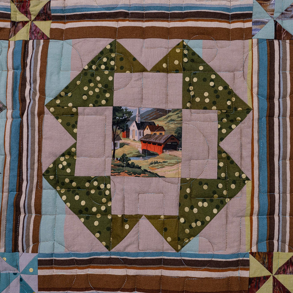 A close-up of a quilted textile featuring a geometric block pattern with a central patch depicting a small farm scene. The design includes olive green polka-dot sections, light blue and white sections, brown and beige stripes, and a blue pattern on the right side.