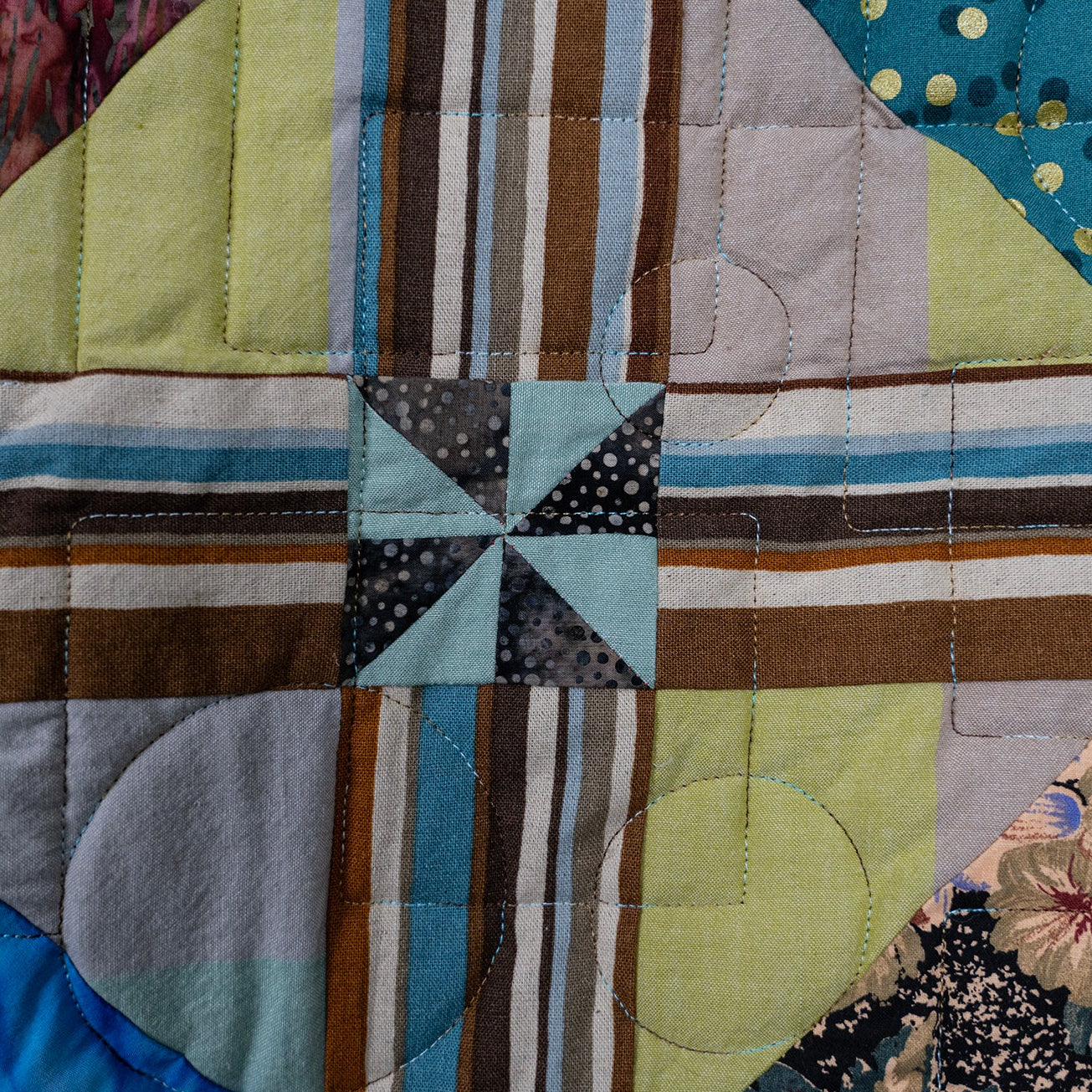 A close-up of a patchwork quilt showing various fabric patterns including stripes, polka dots, floral prints, and solid colors in blue, green, brown, beige, and gray.