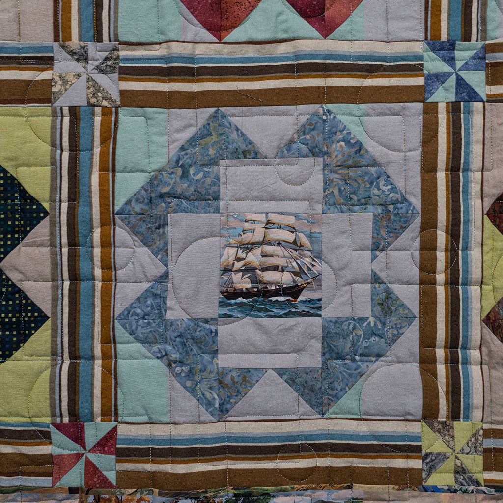 A close-up of a quilted patchwork textile featuring geometric blocks, striped borders, and a central blue ship motif, with other patchwork sections visible around it.