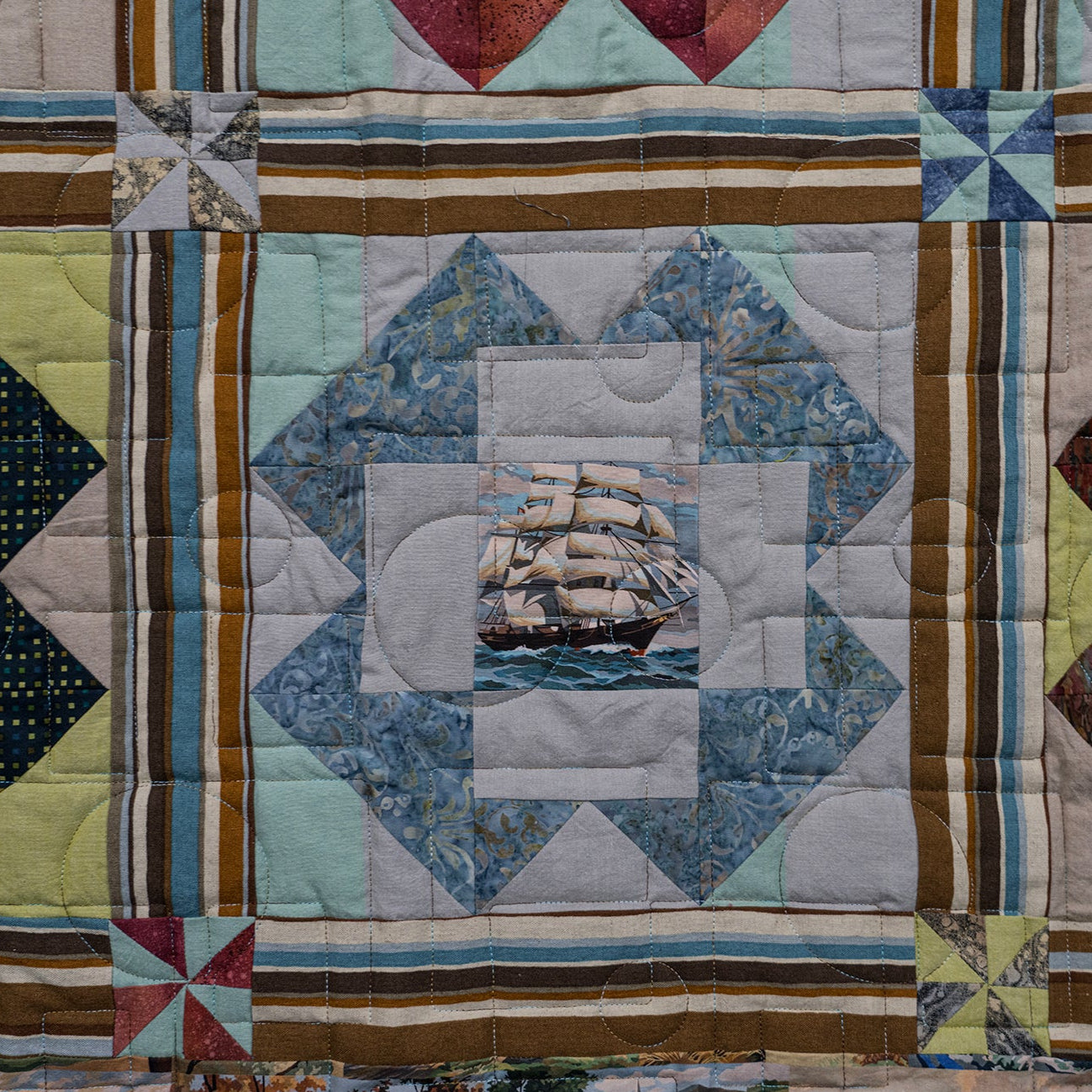 A close-up of a quilted patchwork textile featuring geometric blocks, striped borders, and a central blue ship motif, with other patchwork sections visible around it.