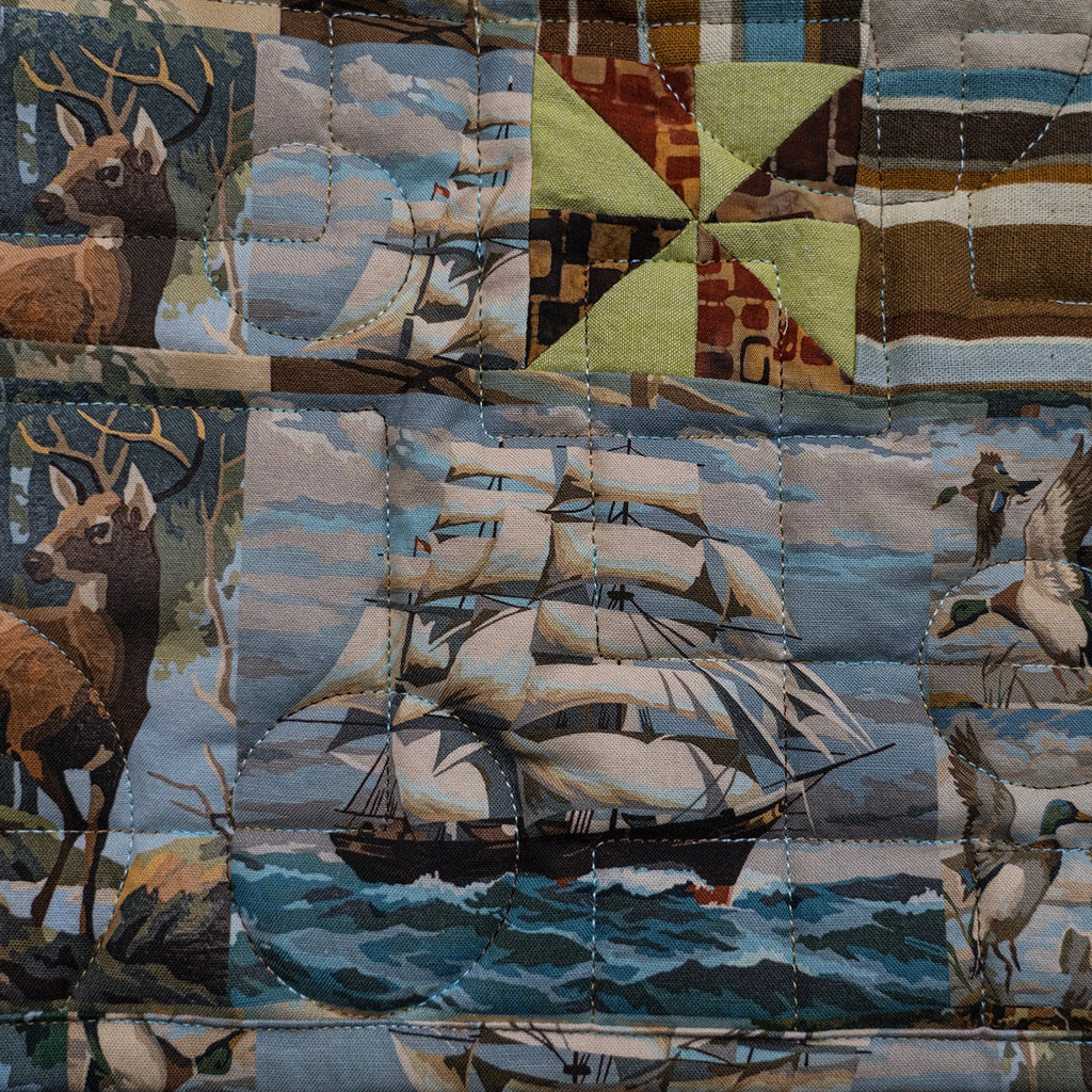 A quilted fabric panel featuring a patchwork design of woodland deer, sailing ships on the water, ducks, and geometric striped blocks in muted earth tones and blues.