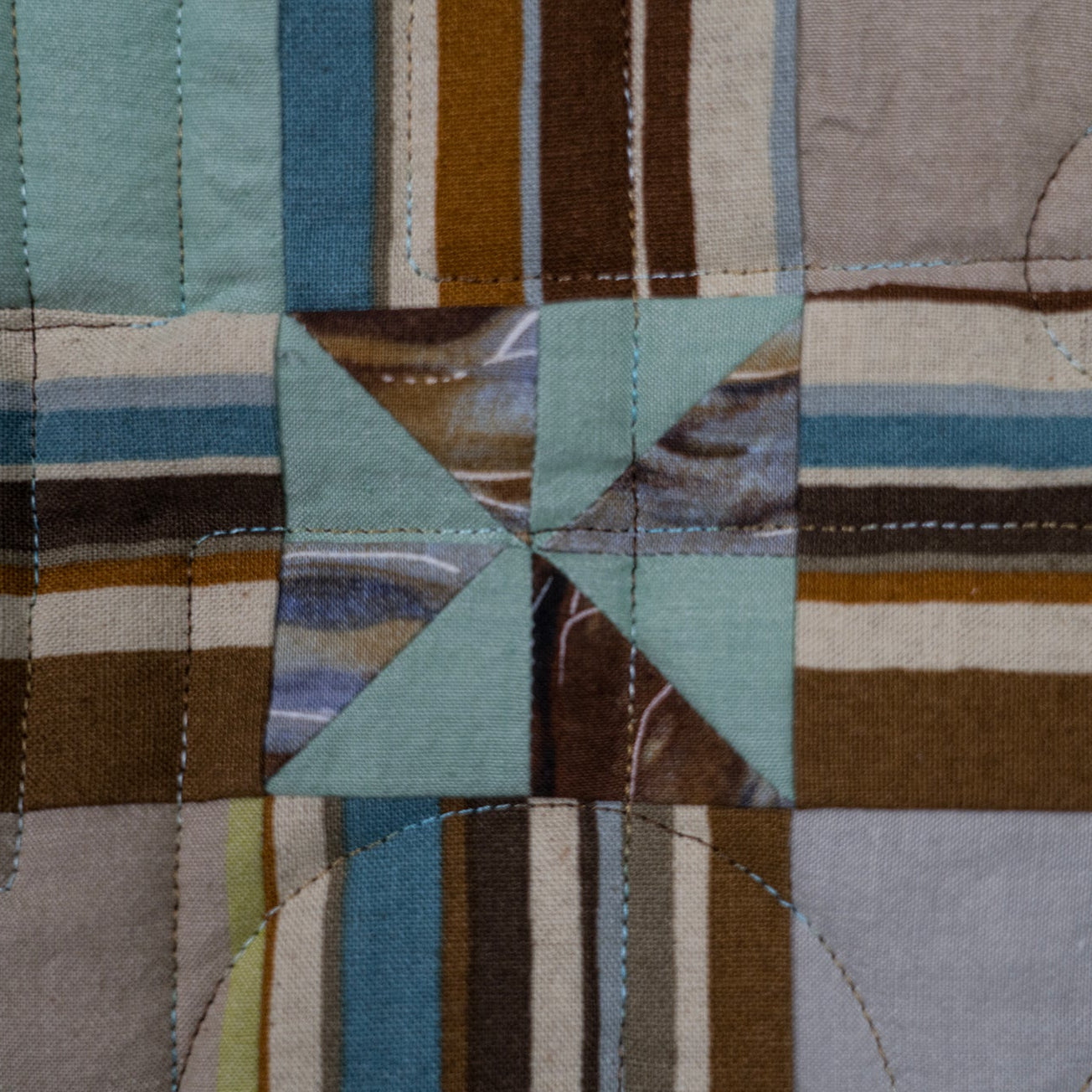 A close-up of a quilted fabric panel featuring a central star patch made of light blue and brown patterned fabric, surrounded by striped and solid blocks in muted blue, brown, beige, and gray.